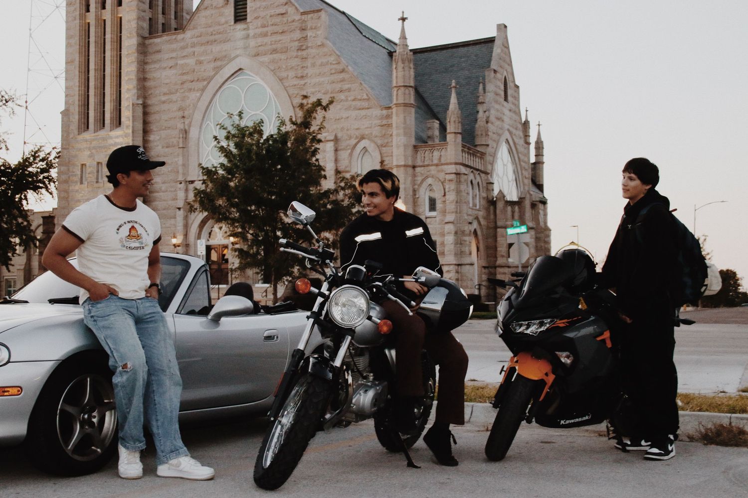Three men gathered around motorcycles on a street corner with a Gothic stone church in the background.