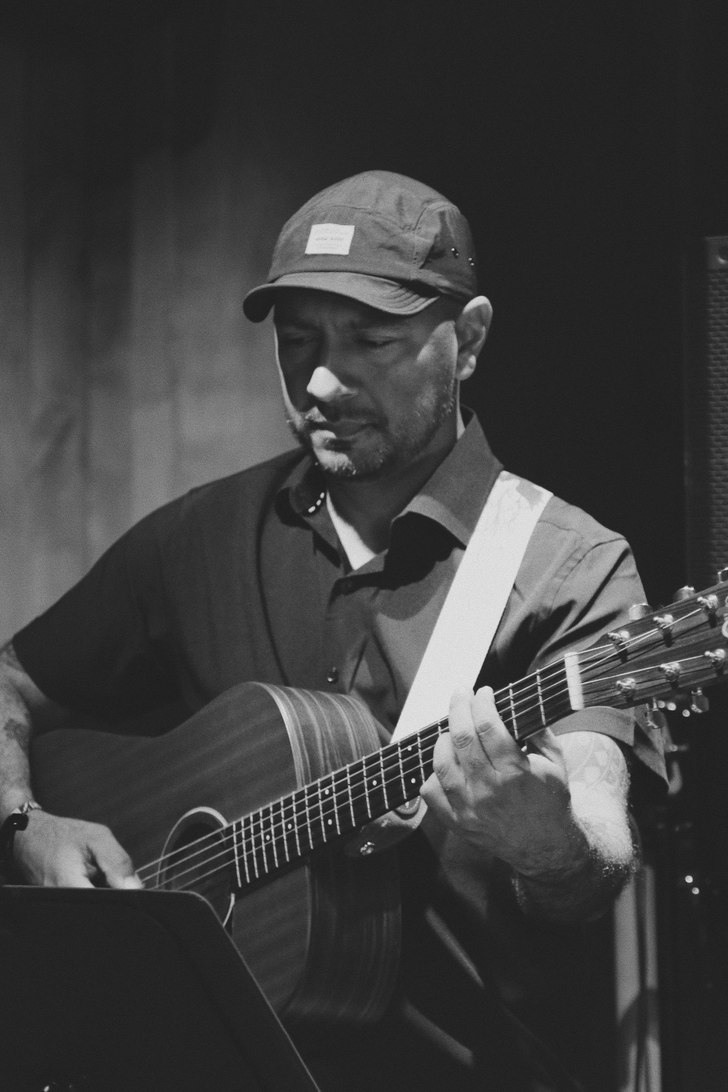 Black and white photo of a man wearing a cap playing acoustic guitar on stage.