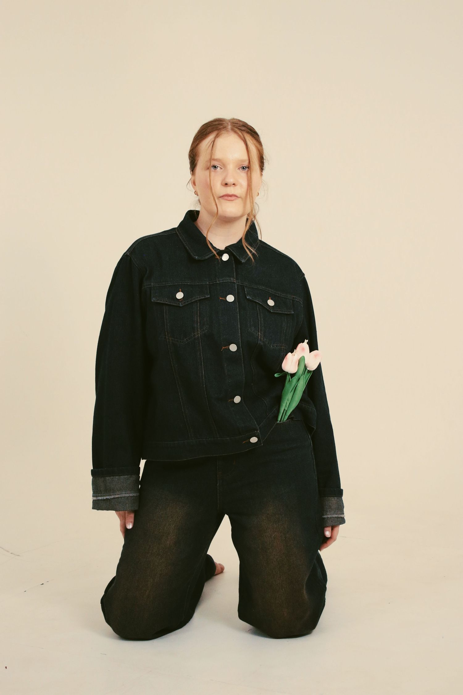 Young woman kneeling in black denim jacket and jeans, holding white tulips, against neutral background.
