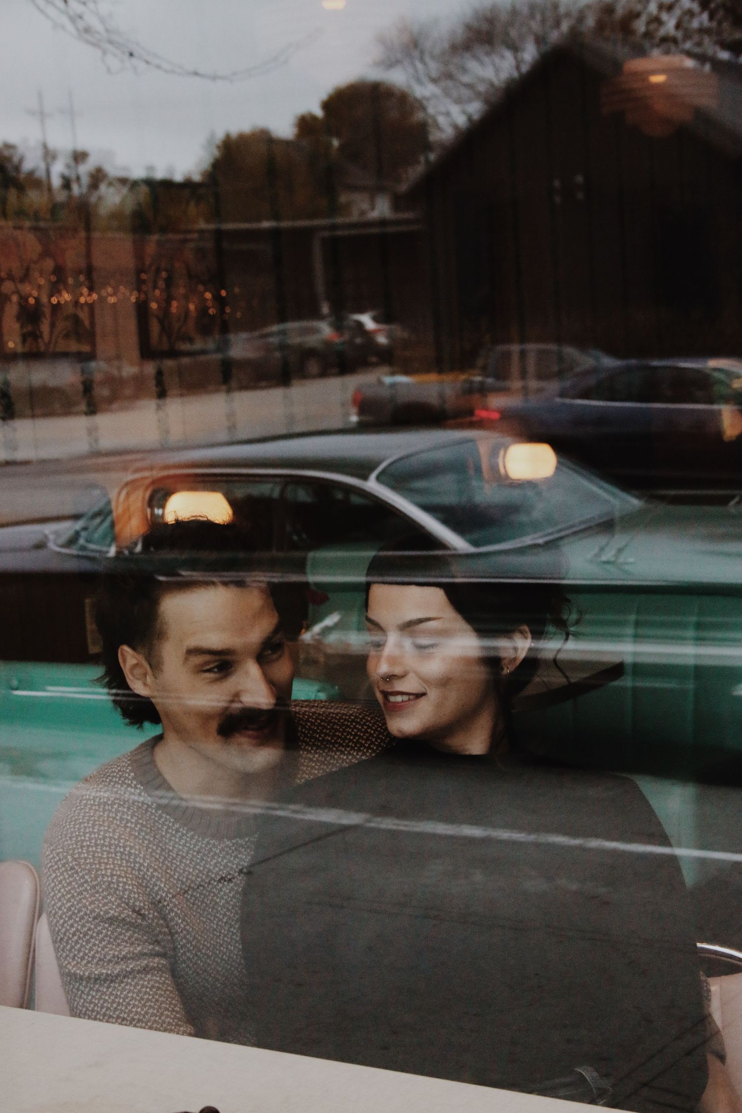 Romantic couple sharing a tender moment photographed through a cafe window with street reflections.
