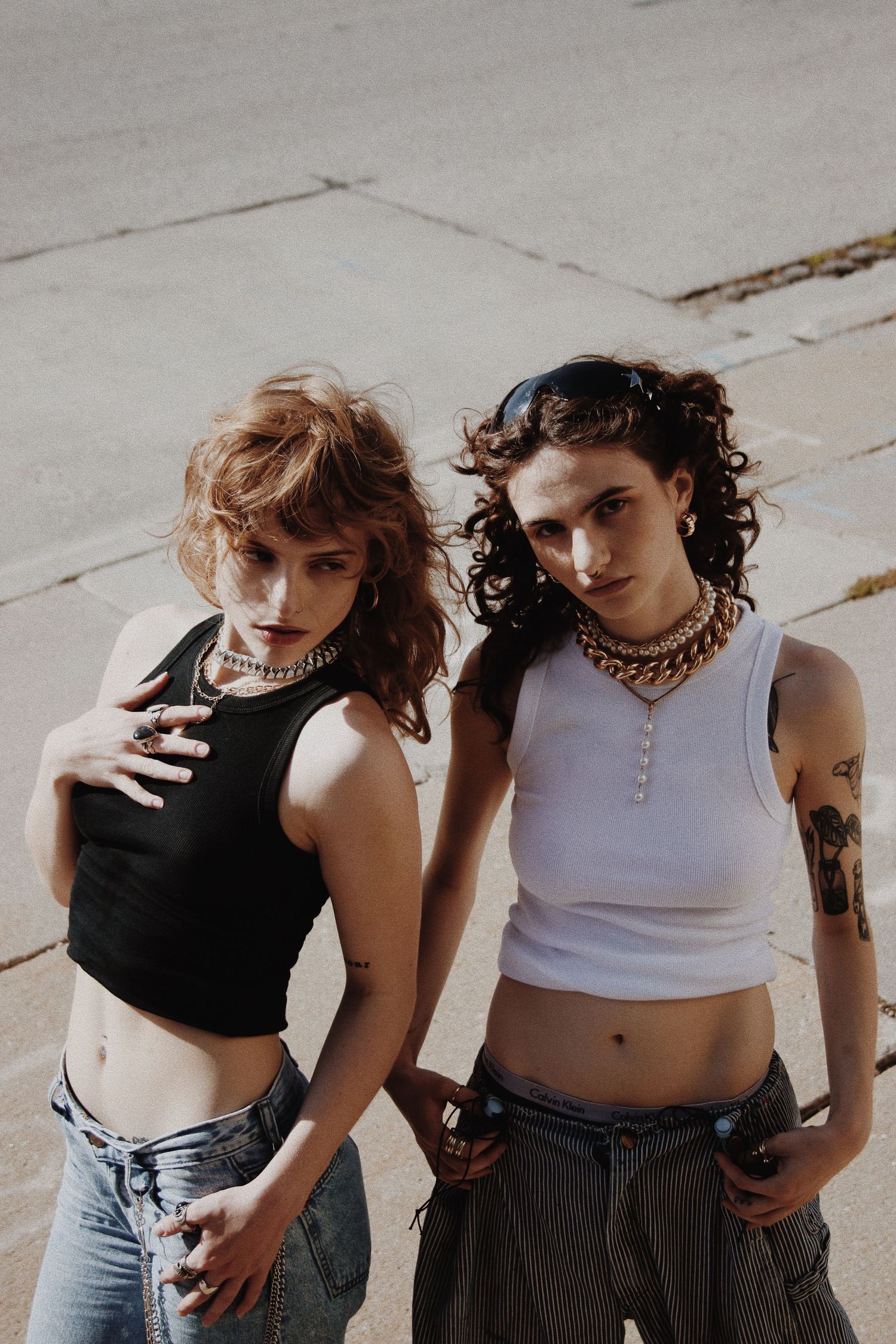 Two young women in crop tops and jeans pose outdoors, wearing gold chain necklaces, one with tattoos.