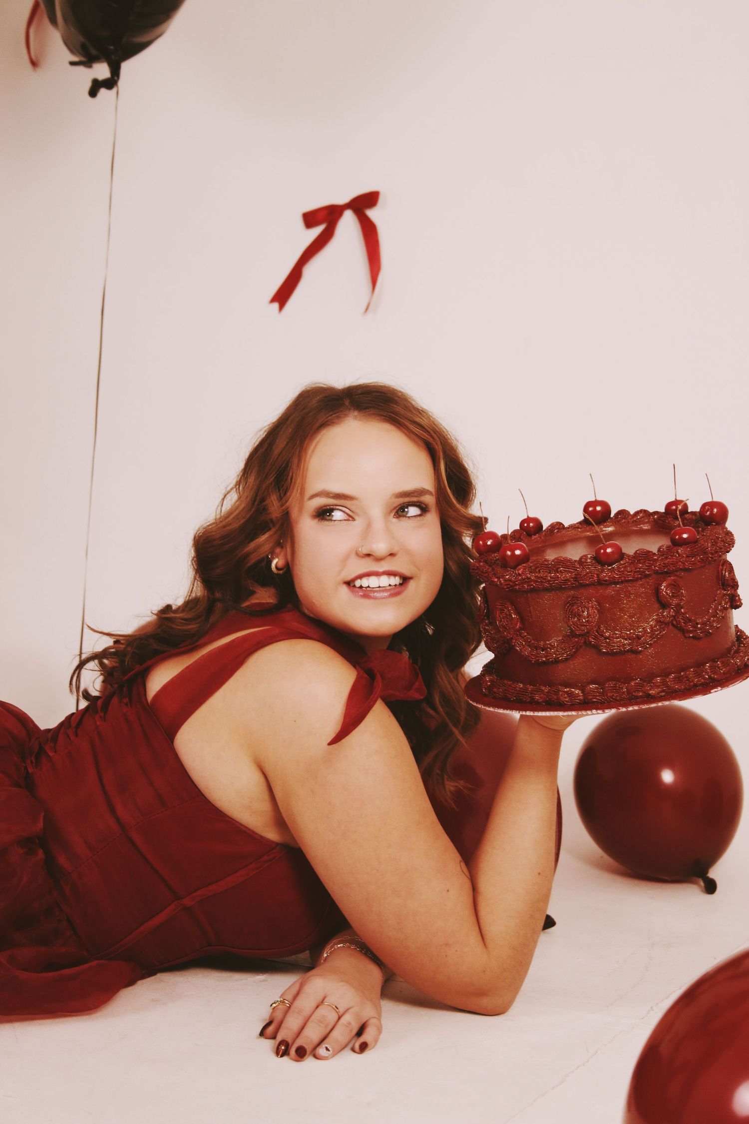 Young woman in red dress lying down smiling at chocolate cherry cake surrounded by red balloons and bow decorations.