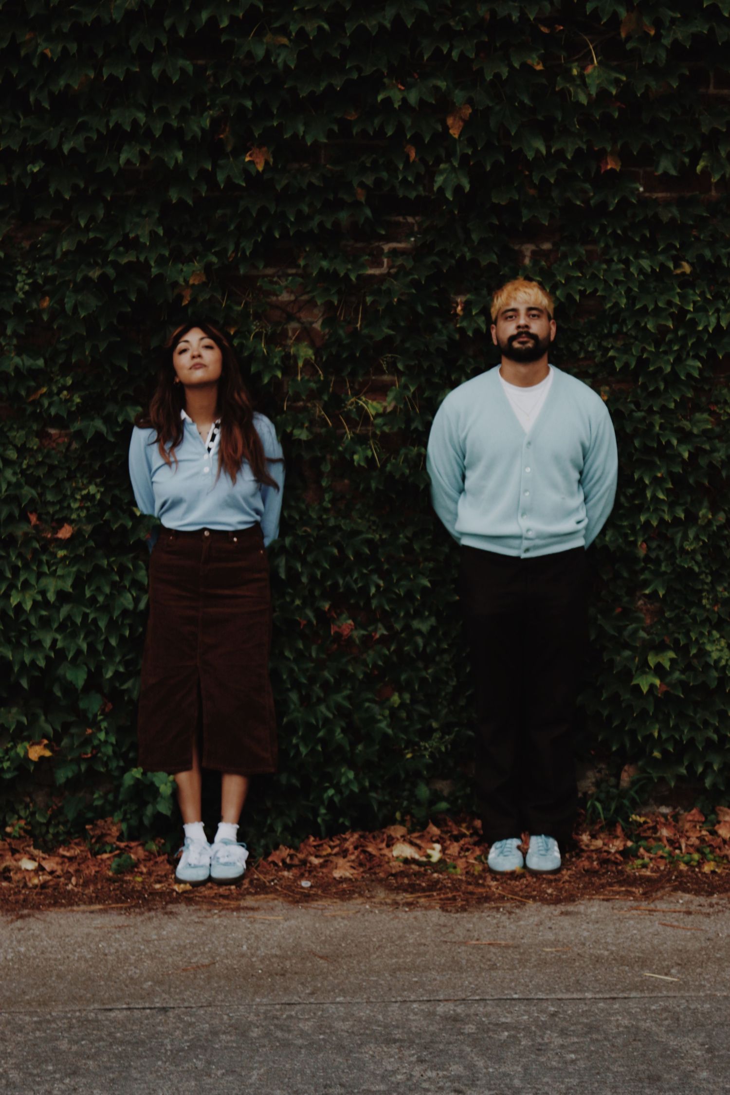 Two young adults stand against an ivy-covered brick wall, wearing casual blue and brown outfits, on a sidewalk.