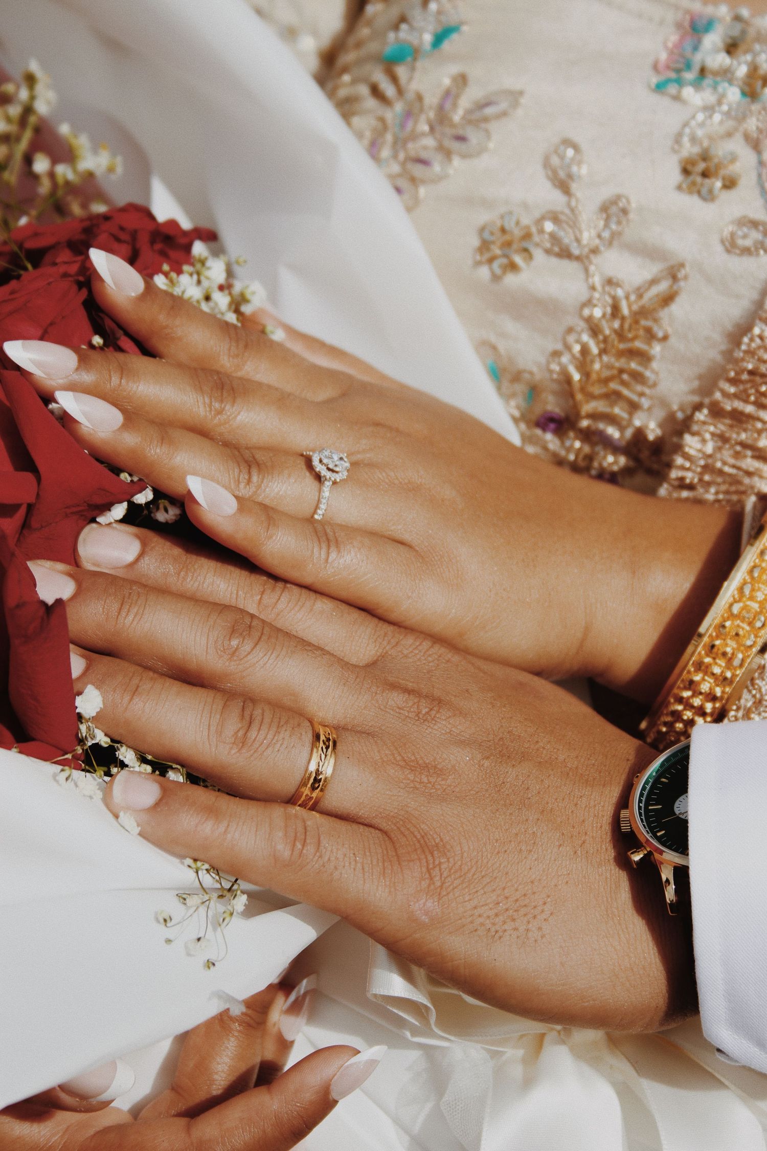 Close-up of bride and groom hands showing diamond engagement ring and gold wedding bands with bouquet.
