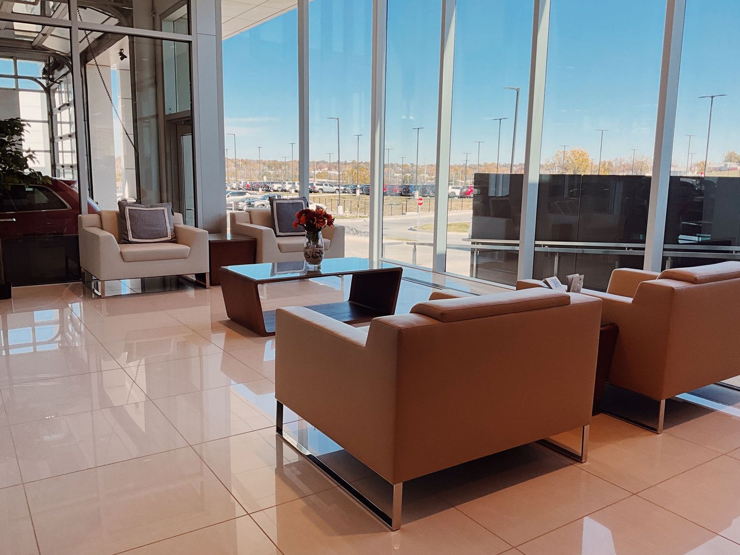 Modern car dealership waiting lounge with tan leather chairs, large windows, TV, and glossy tile floors.
