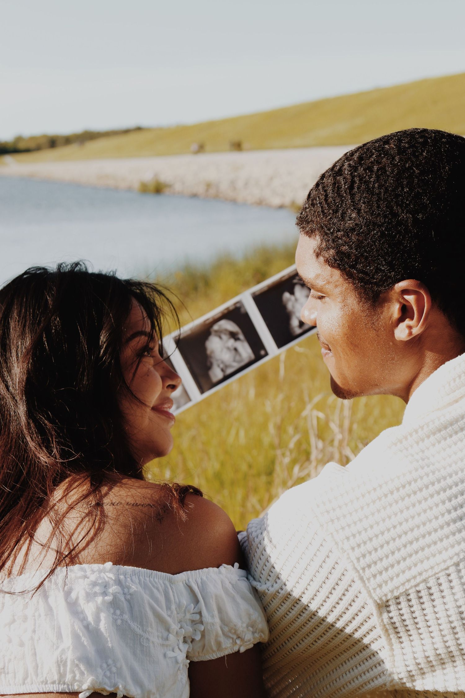 Happy couple smiling at each other while holding ultrasound photos outdoors near a lake, announcing pregnancy.