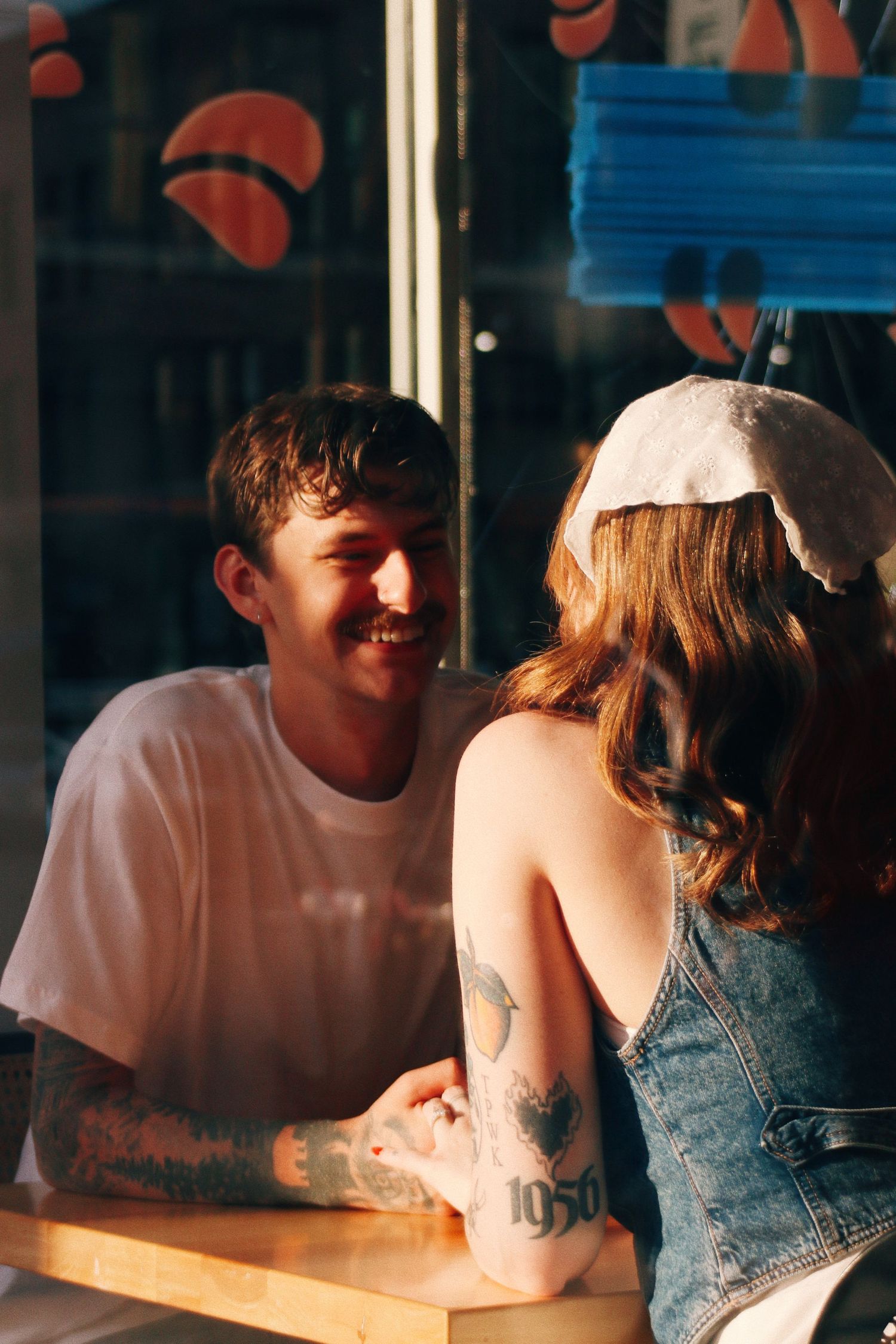 A smiling tattooed couple holding hands at a restaurant table, bathed in warm golden sunlight.