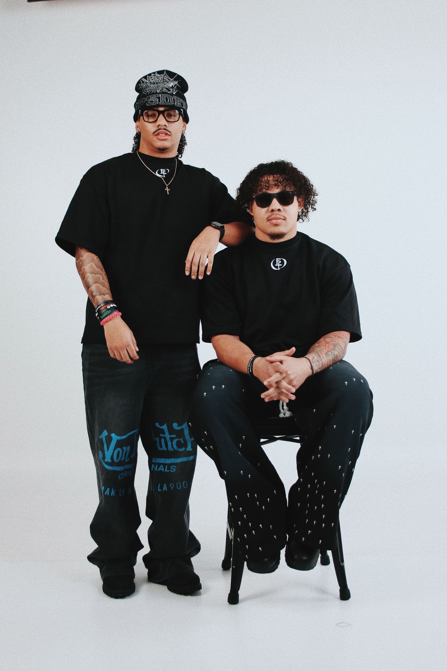 Two young men in matching black outfits and sunglasses pose in a studio, one standing and one seated on a stool.