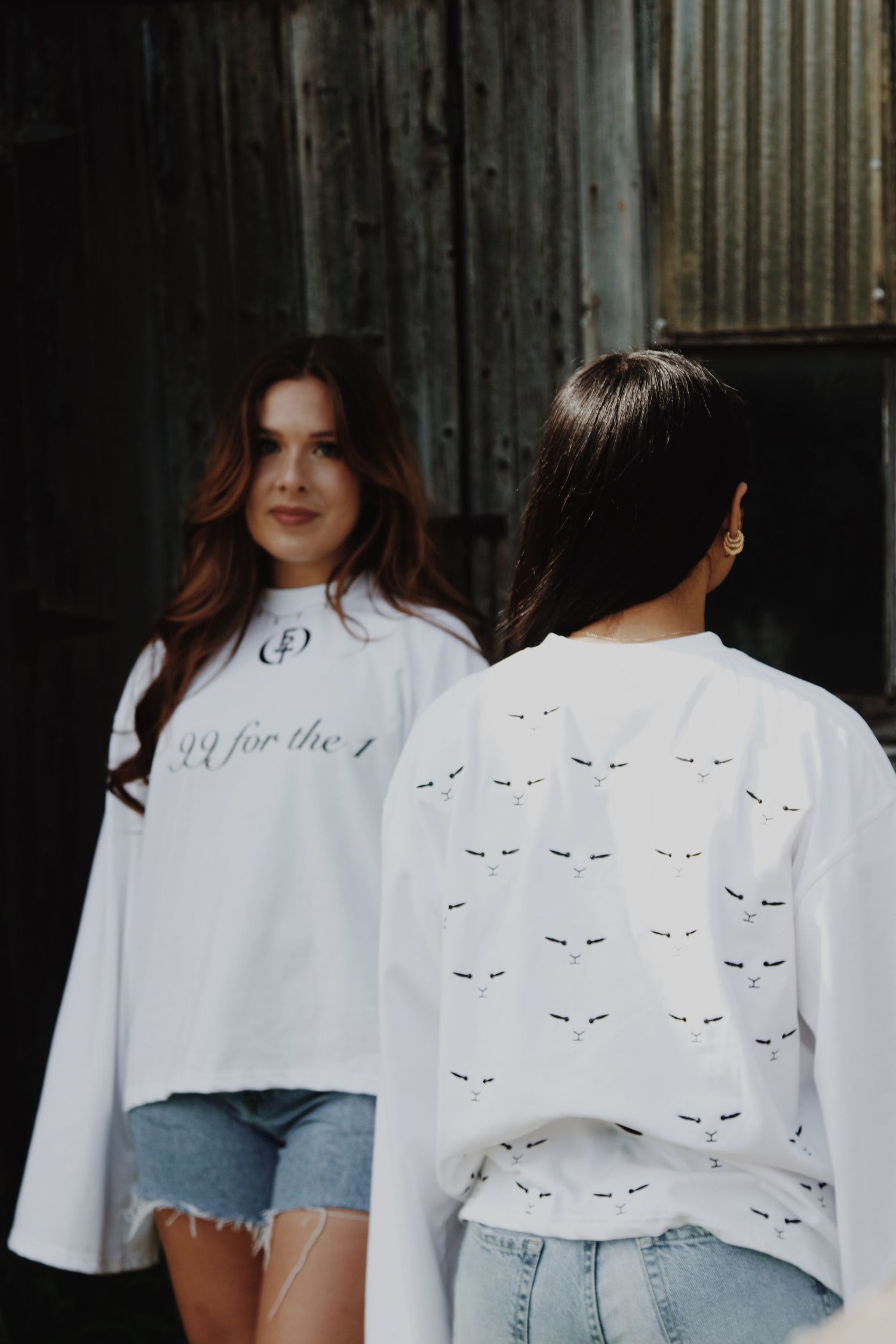 Two women modeling white oversized long-sleeve shirts with small graphic prints, standing near a weathered wooden structure.