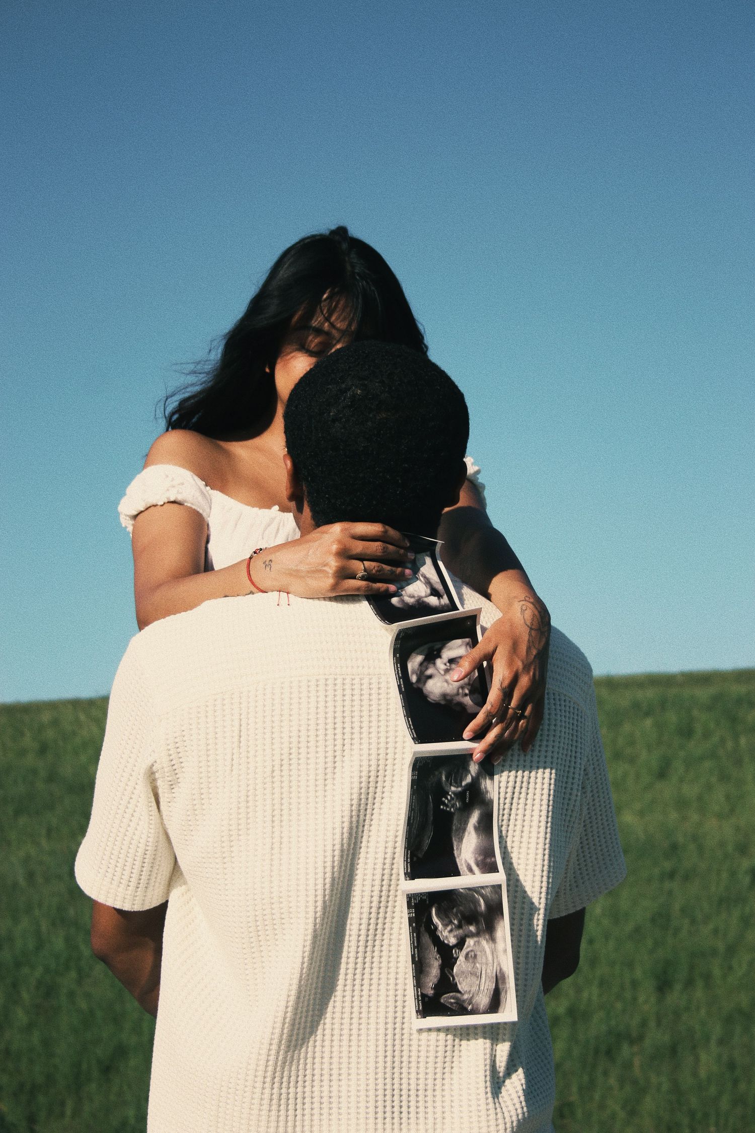 Couple embracing in green field holding ultrasound photos, pregnancy announcement outdoors under blue sky.