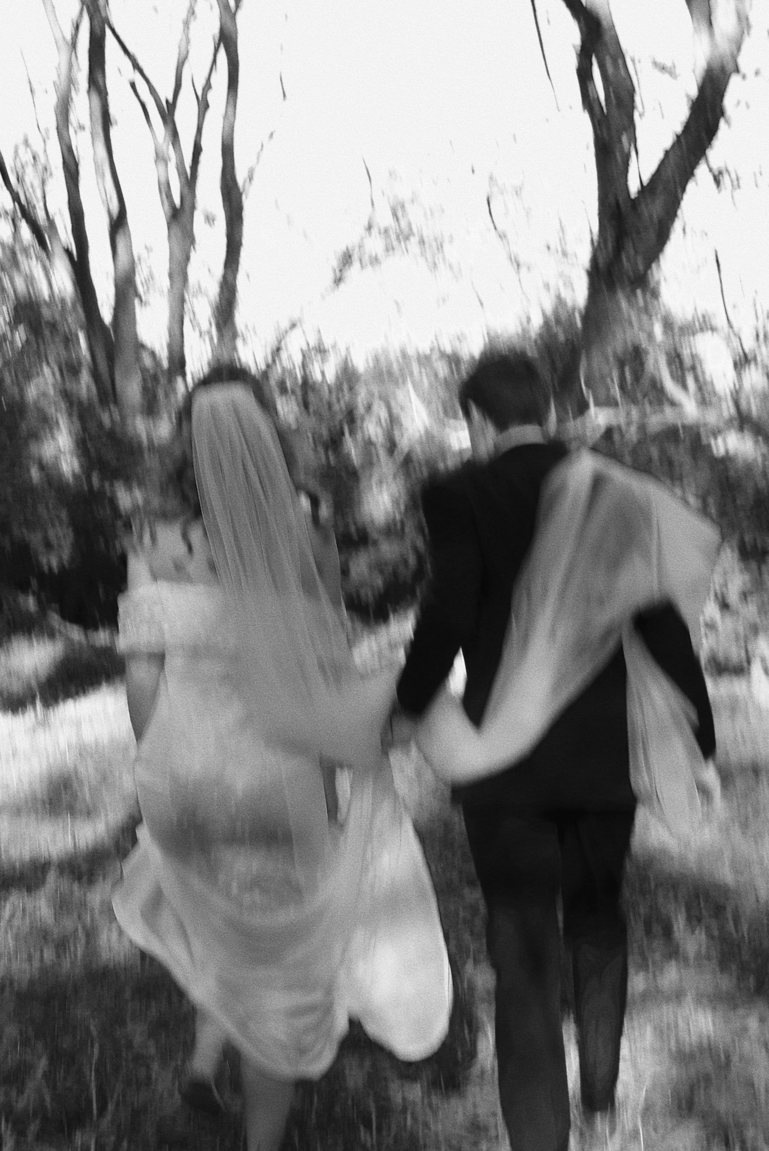 Blurry black and white photo of a bride and groom walking hand in hand through a tree-lined path.