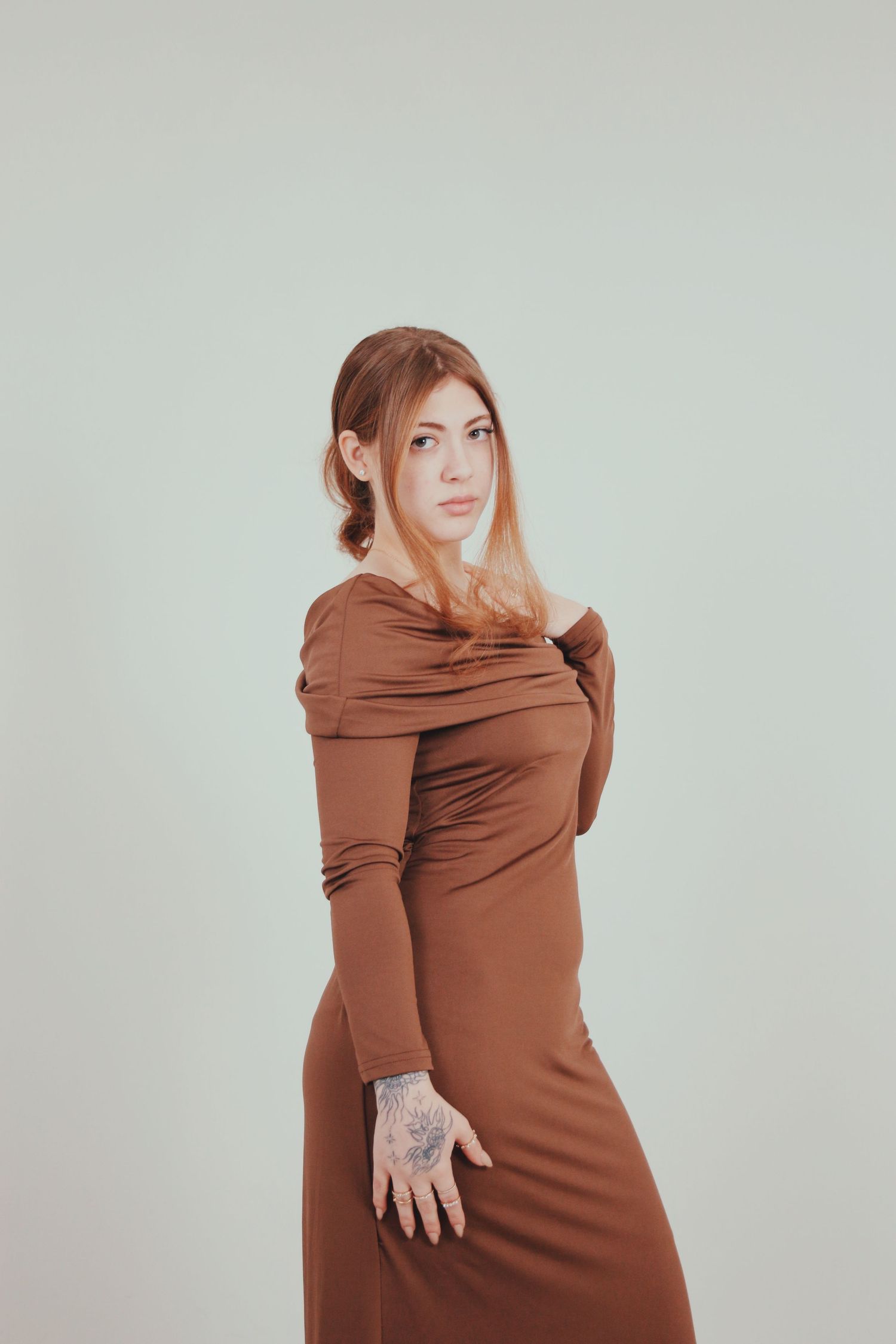 Woman in fitted brown off-shoulder long sleeve dress posing with hand tattoo against light background.