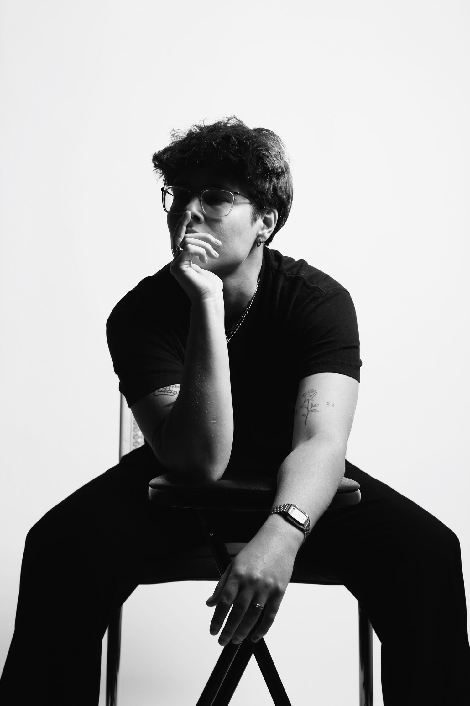 Black and white portrait of a young man in glasses sitting on a chair, wearing all black with a watch and necklace.