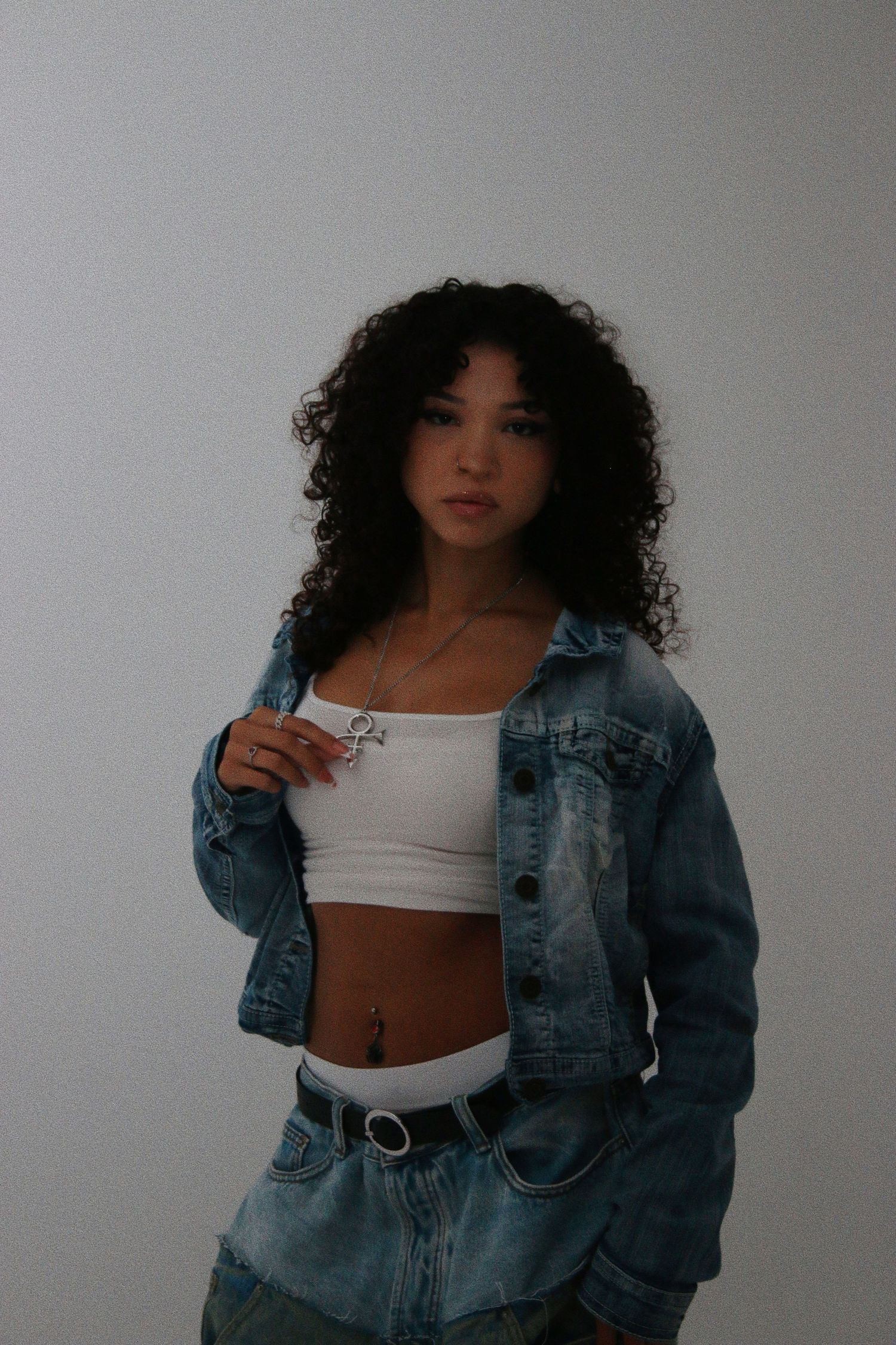 Young woman with curly hair wearing a white crop top, denim jacket, and jeans poses against a white wall.