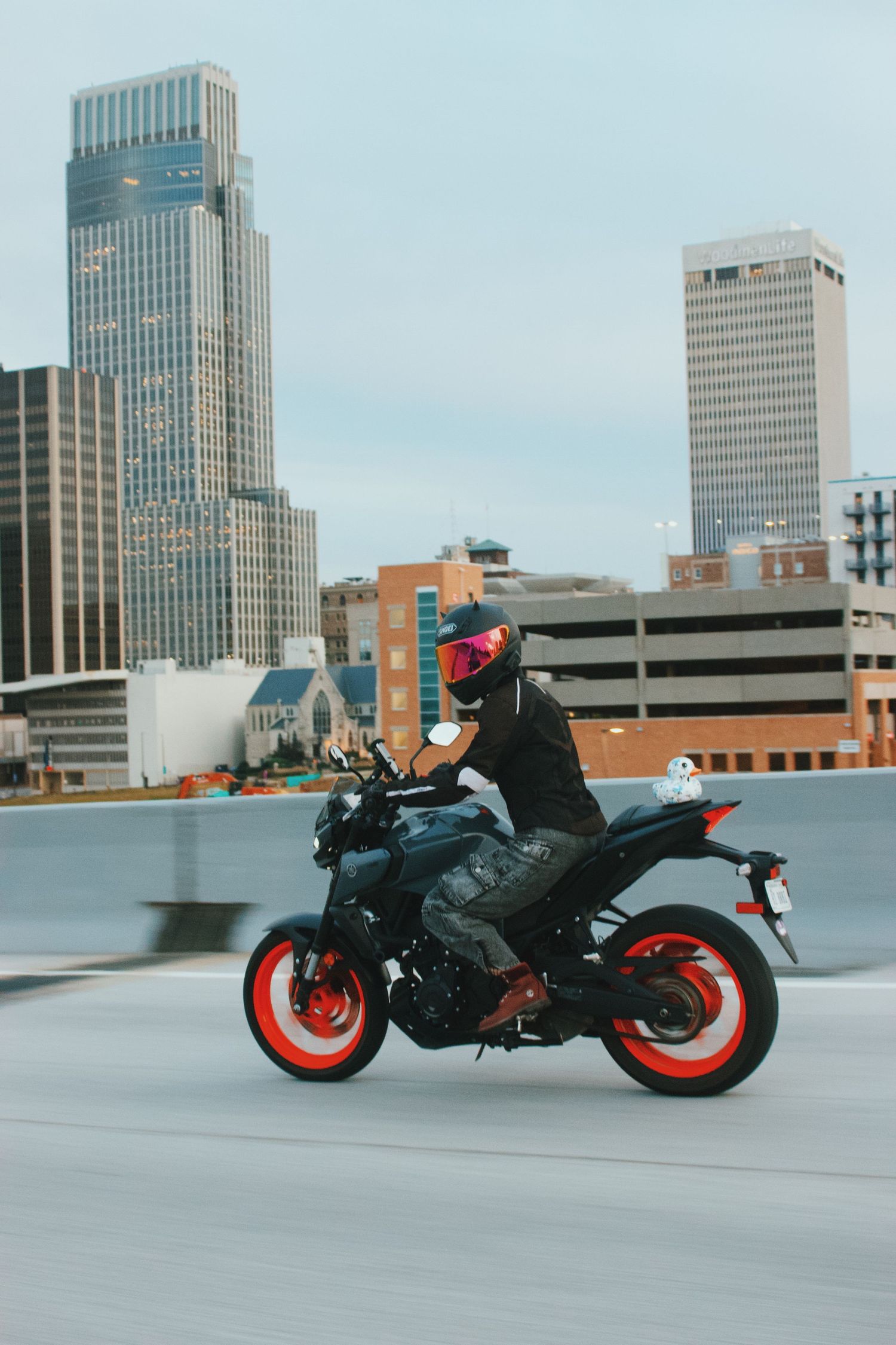 Motorcyclist in black jacket and iridescent helmet rides a black and red Yamaha MT on urban highway with city skyline.
