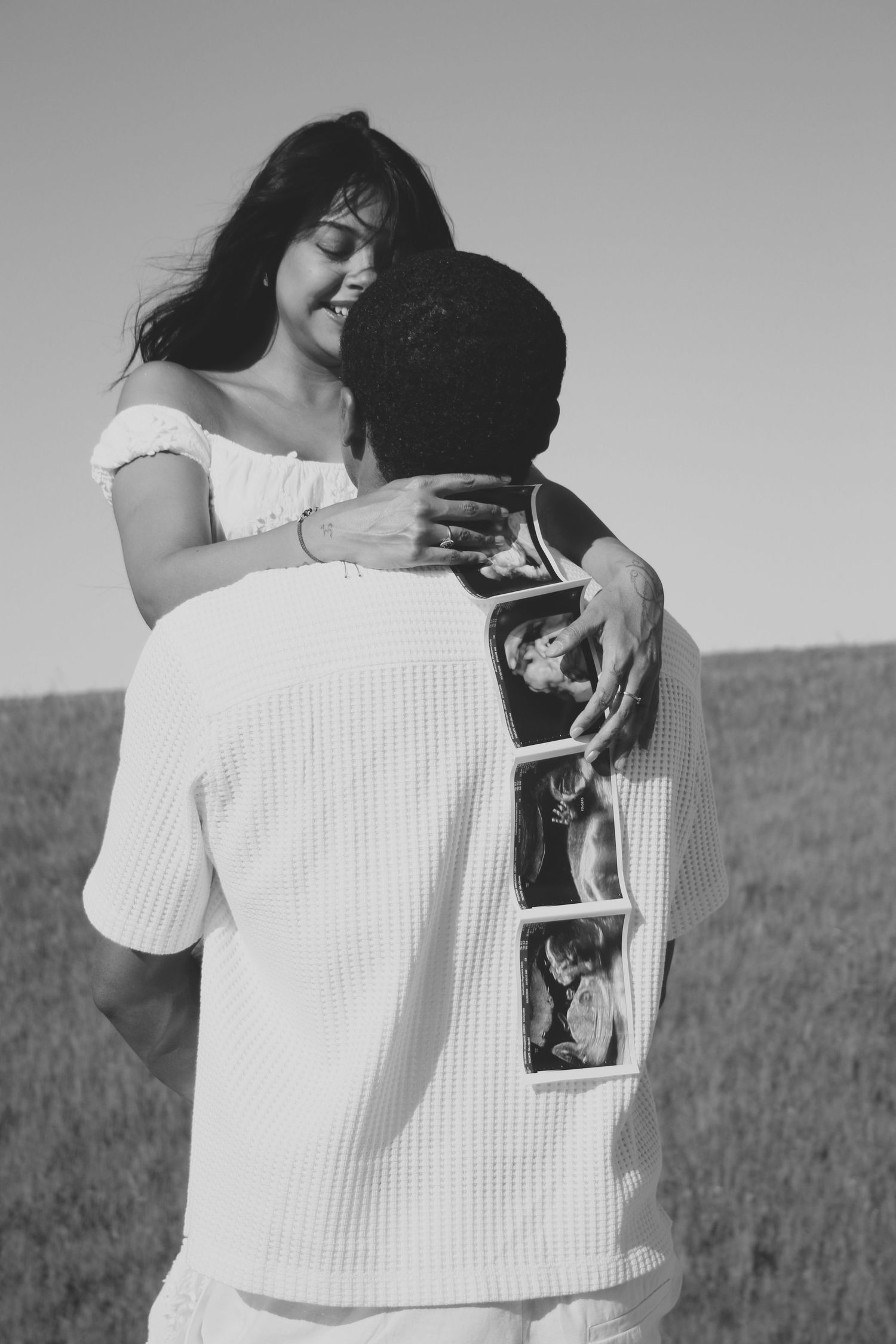 Black and white photo of a couple embracing outdoors, woman smiling joyfully, man holding ultrasound photos on his back.