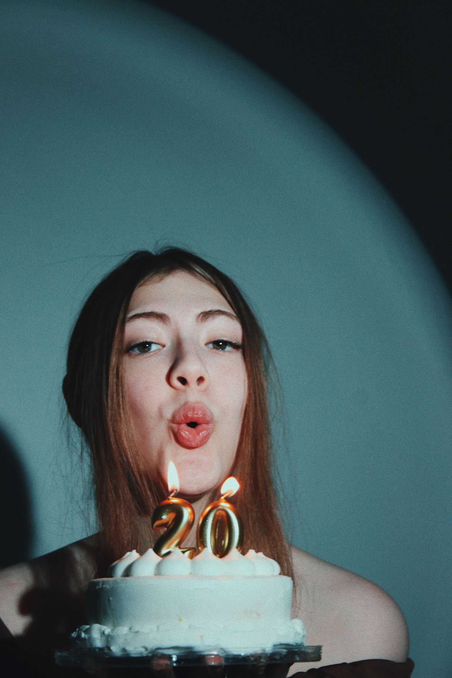 Young woman blowing out golden number 20 candles on a white birthday cake in dim lighting.