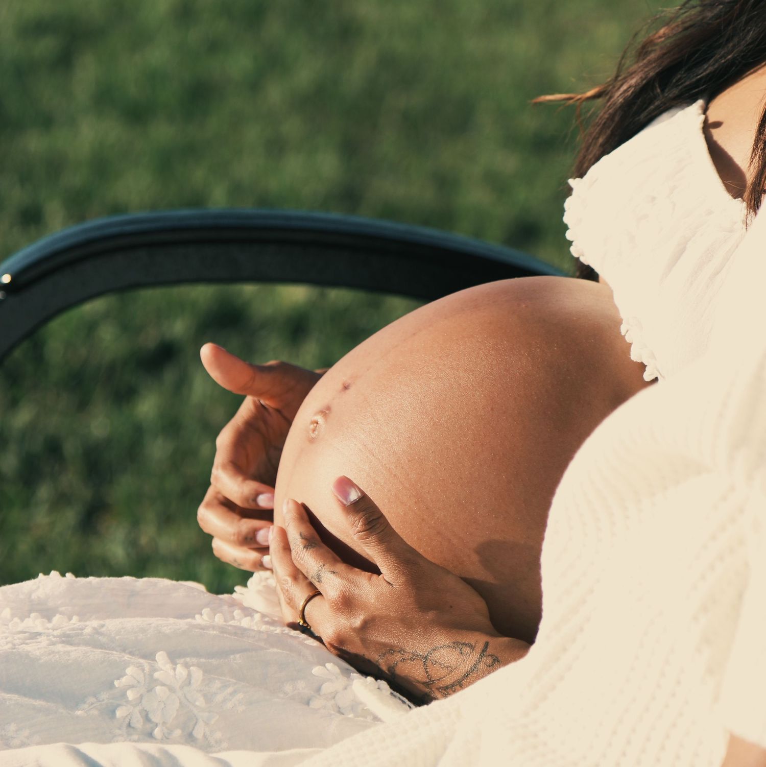 Pregnant woman in white dress cradling her bare baby bump outdoors on a sunny day.