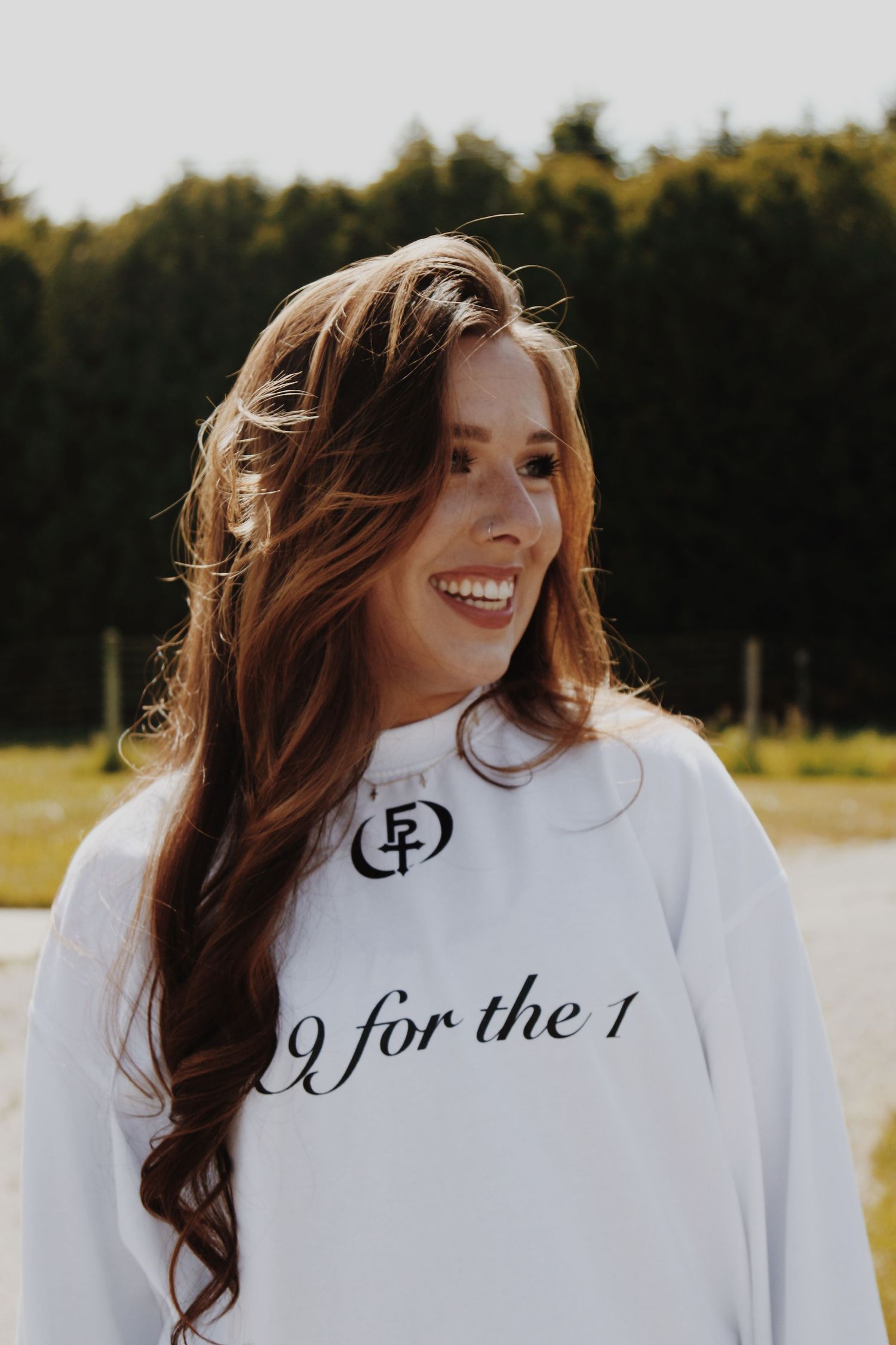 Smiling young woman with long auburn hair wearing a white sweatshirt outdoors near a forest.