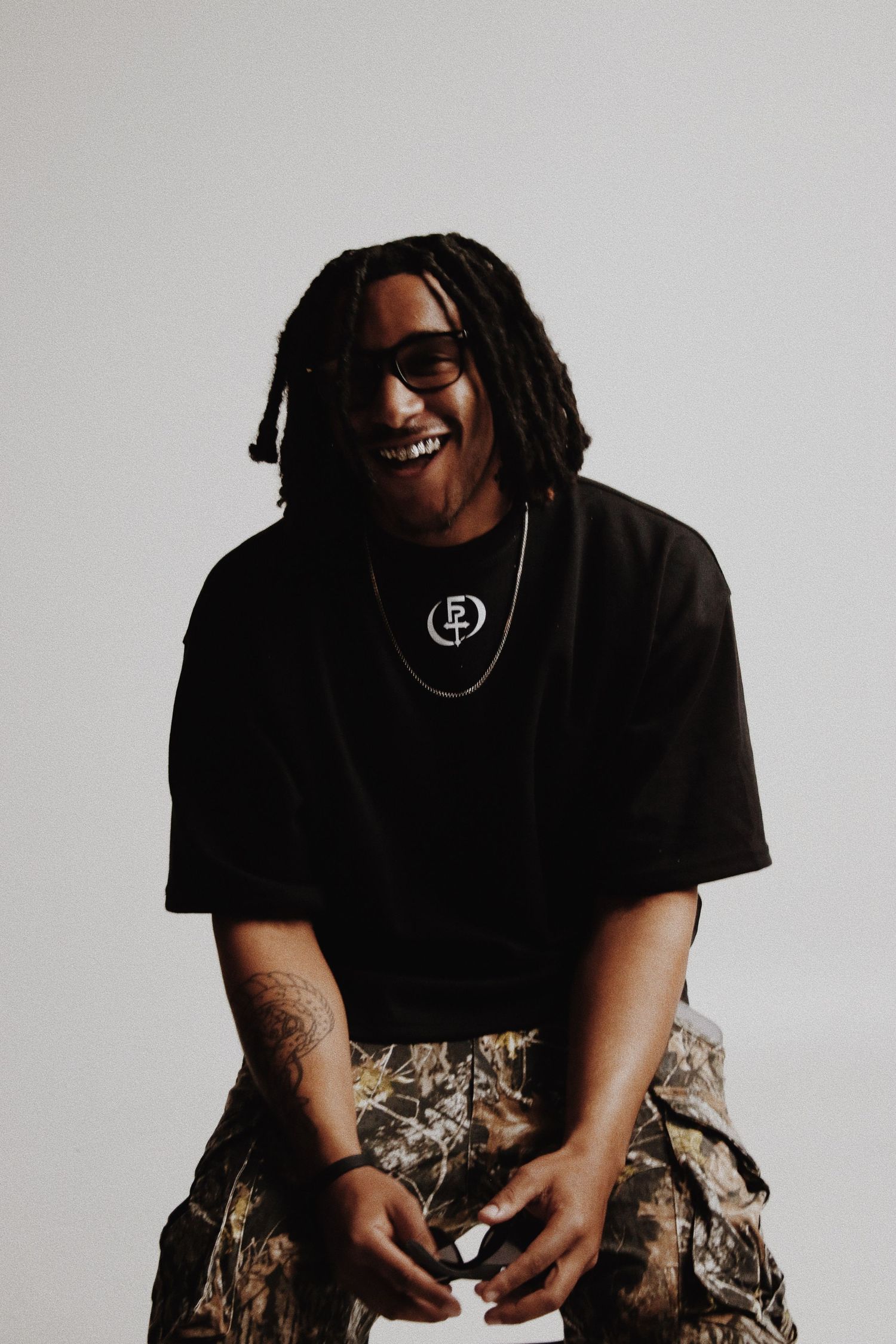 Smiling man with dreadlocks wearing black graphic tee, chain necklace, camo pants, and sunglasses against gray background.