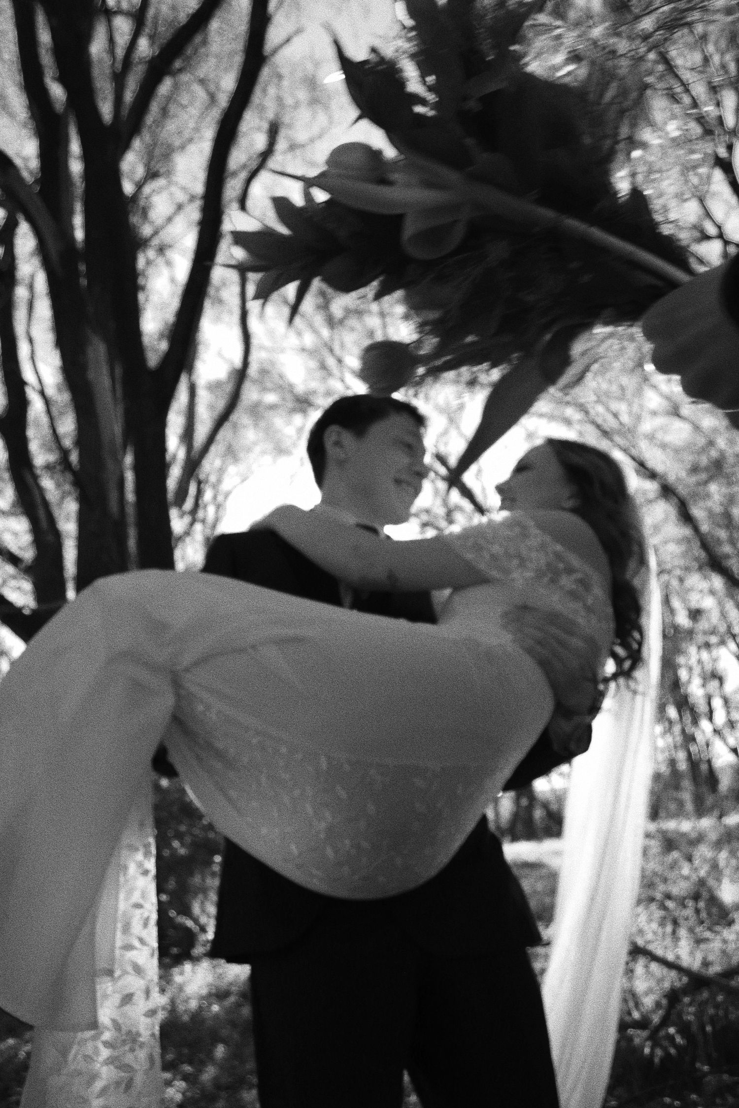 Black and white photo of groom lifting bride in wedding dress outdoors under floral arch surrounded by trees.