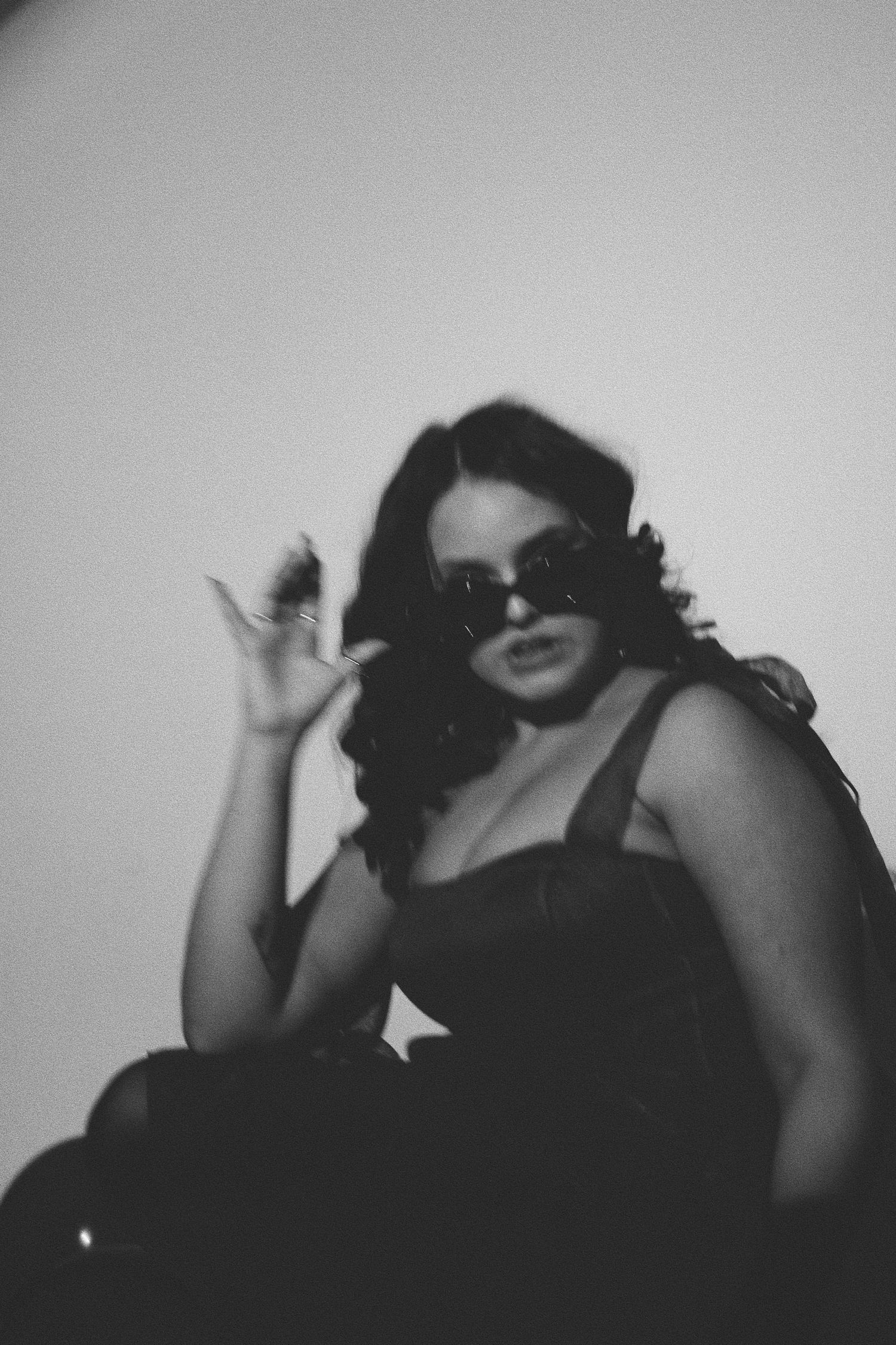 Black and white portrait of a woman in sunglasses and dark dress, posing with hand raised in moody lighting.
