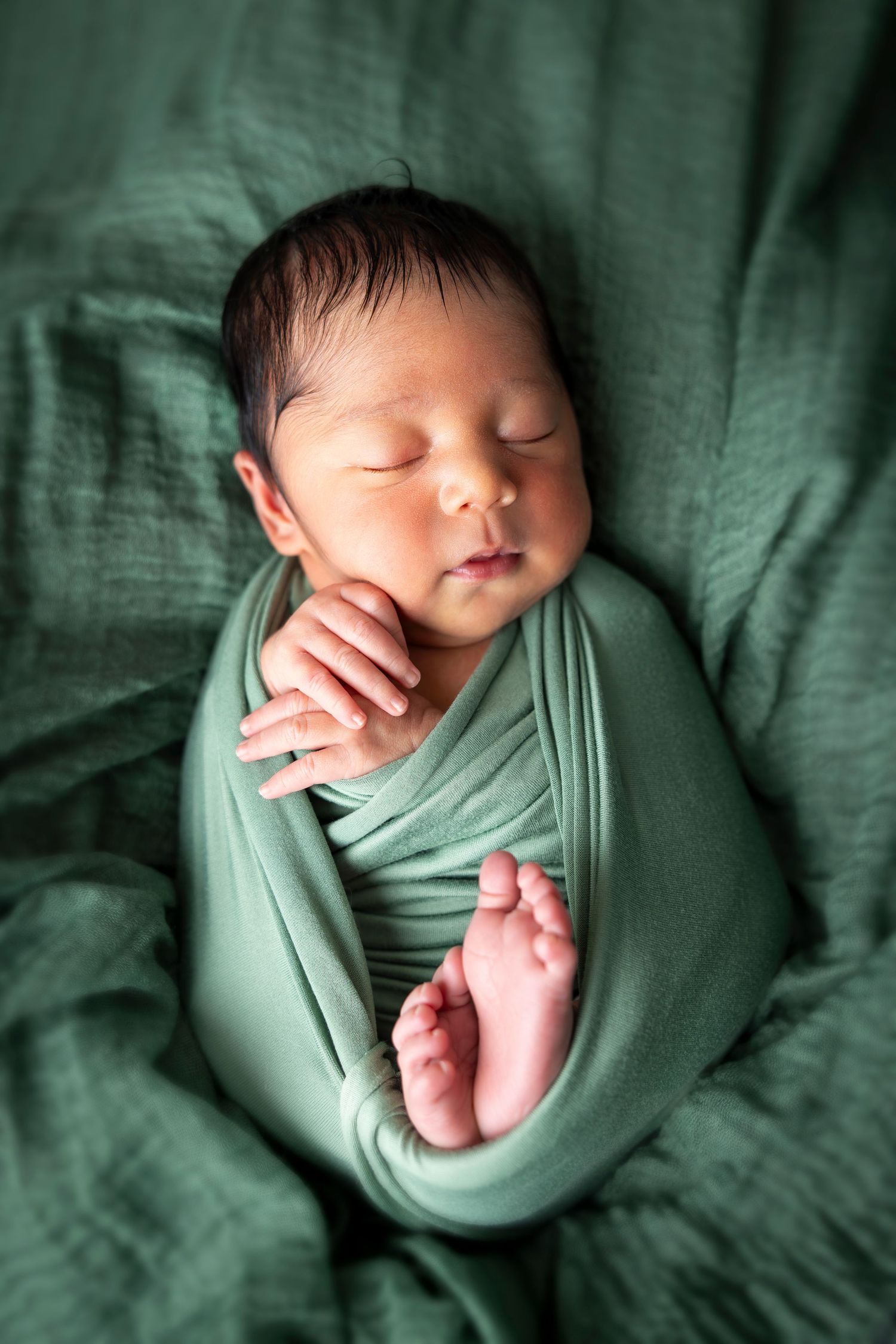 Close-up of a sleeping newborn with dark hair swaddled in a green wrap with tiny feet showing.