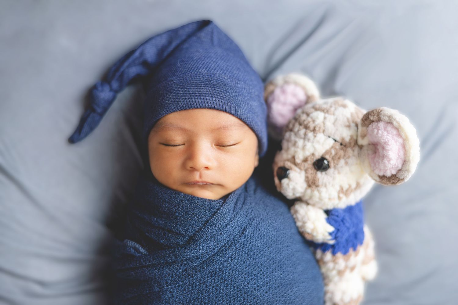 Sleeping newborn in a blue knotted hat and swaddle lying beside a cute crocheted mouse stuffed toy on a soft gray-blue blanket.
