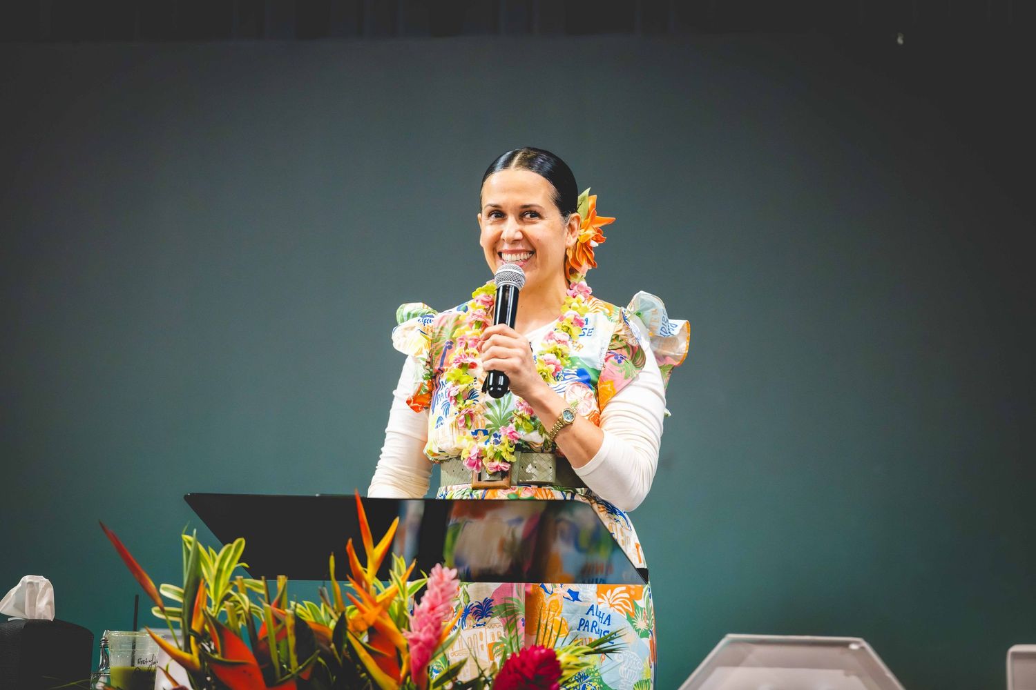 Smiling woman in a floral dress and flower hair accessory speaks into a microphone at a podium with colorful tropical flowers.