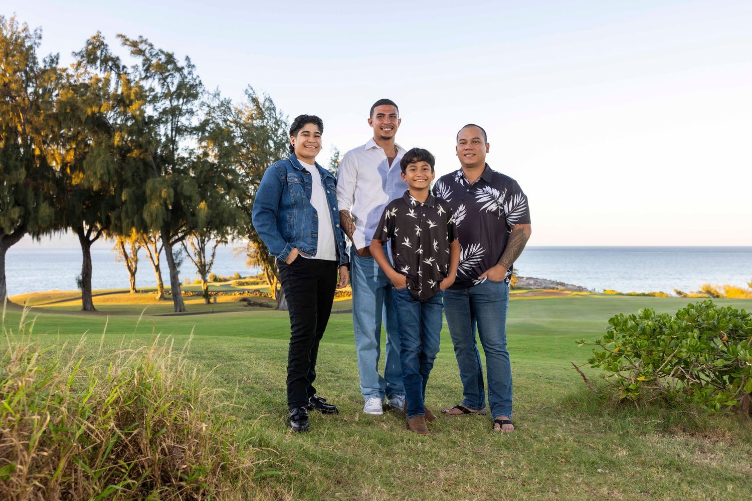 Four people in casual clothing pose together on a grassy coastal lawn with trees and the ocean behind them under a clear sky.