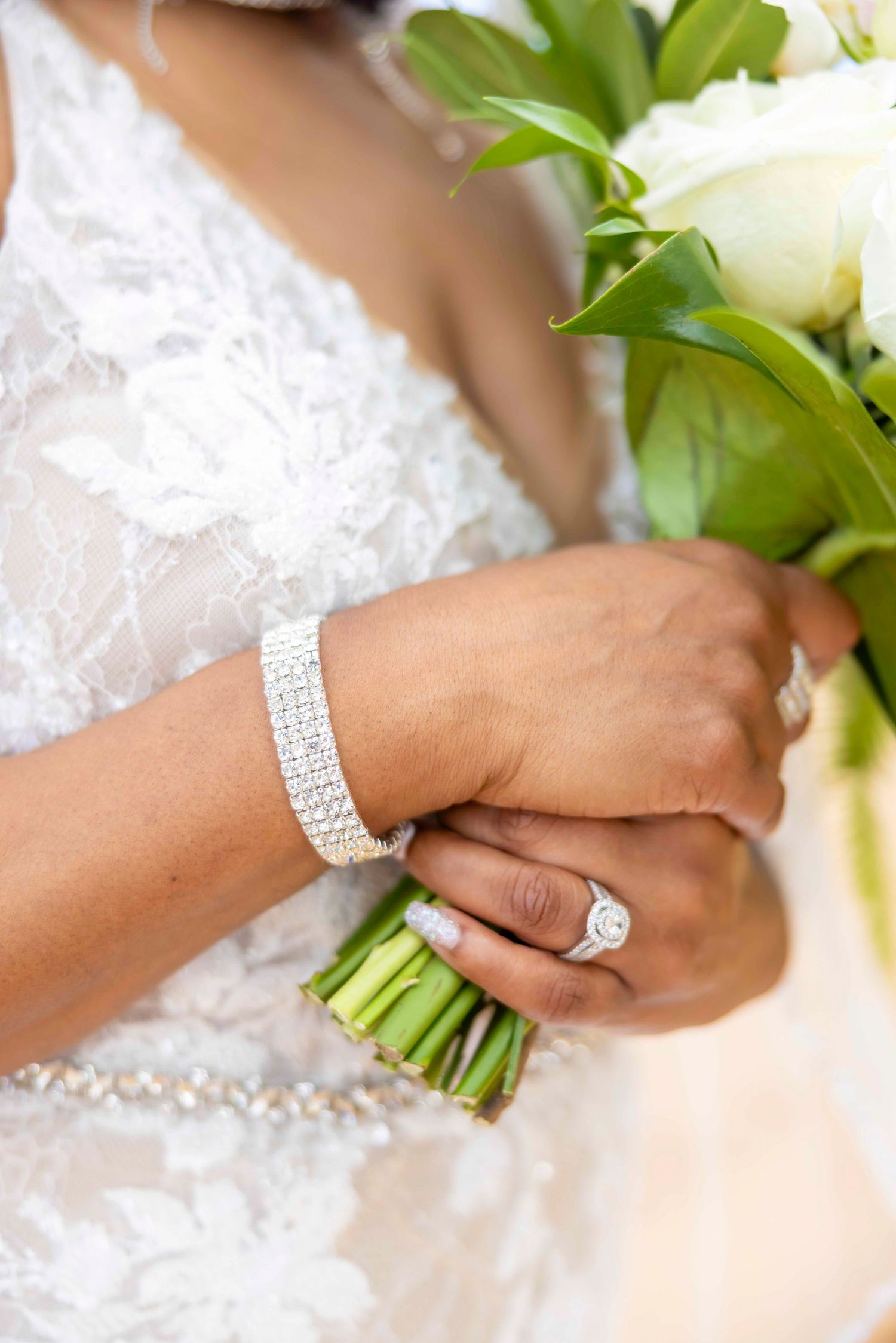 Bride in a white lace wedding gown holds a bouquet of white roses, showing a halo diamond engagement ring and a sparkling bracelet. Wailea