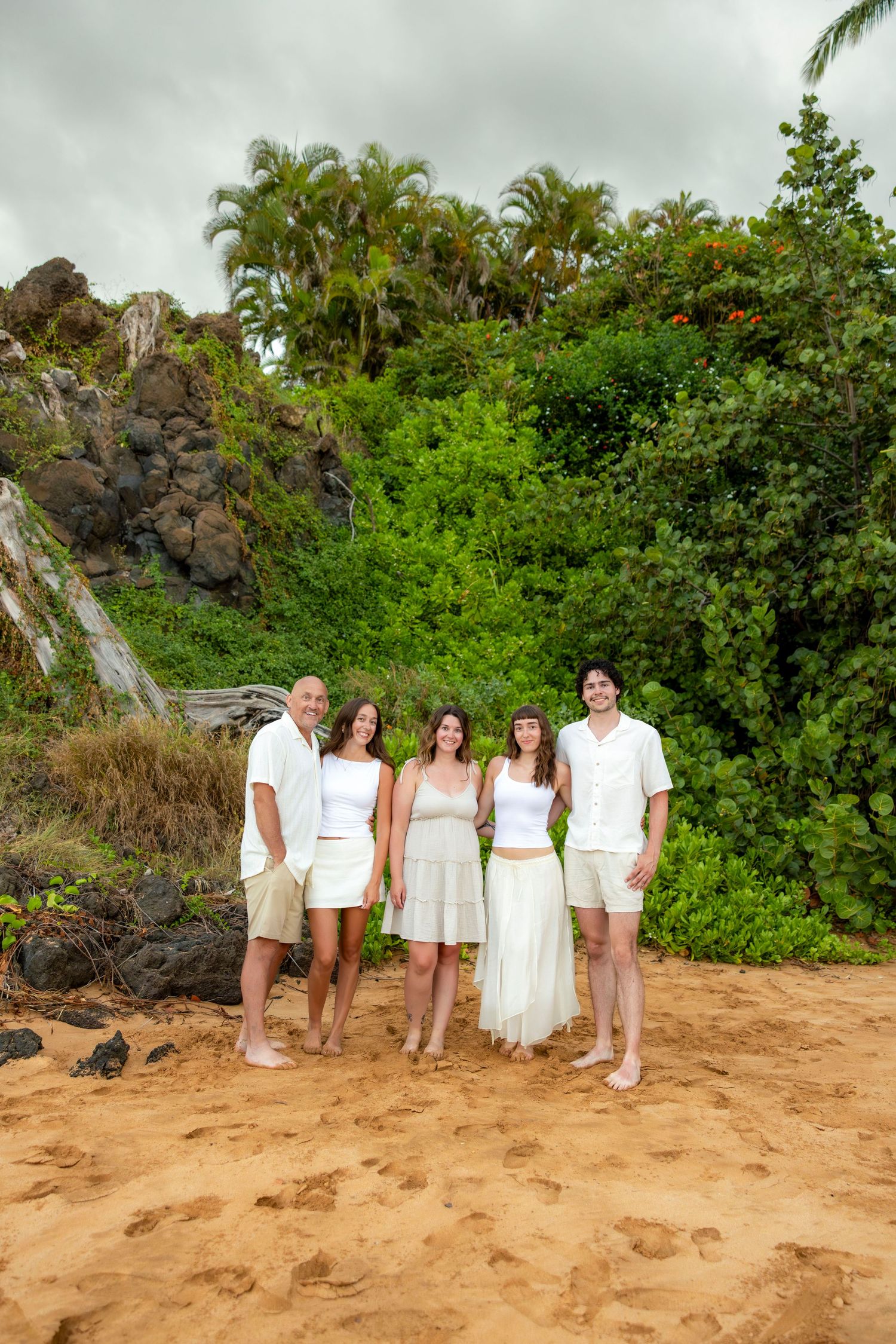 Family of five dressed in white standing together on a golden sandy beach with lush tropical greenery and palm trees behind them.