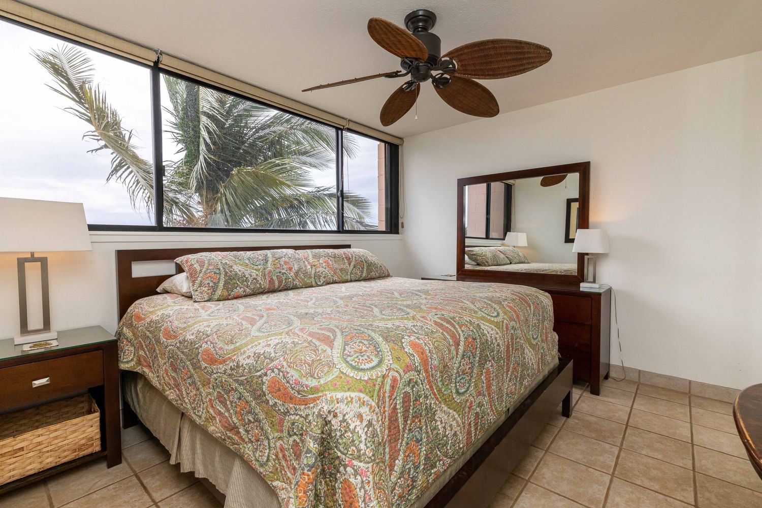 Tropical bedroom with a paisley-covered bed, ceiling fan, wooden nightstands, tile floor, and a large window showing palm trees.