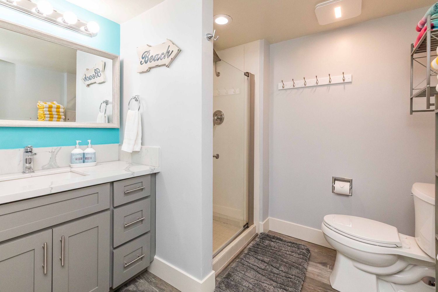 Bright bathroom with gray vanity, teal accent wall, glass-door walk-in shower, white toilet, and wall hooks above a gray patterned rug.