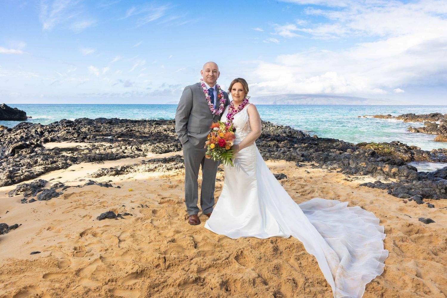 Smiling couple in wedding attire with leis and a bouquet pose on a sandy Hawaii beach with lava rocks and ocean behind. Makena