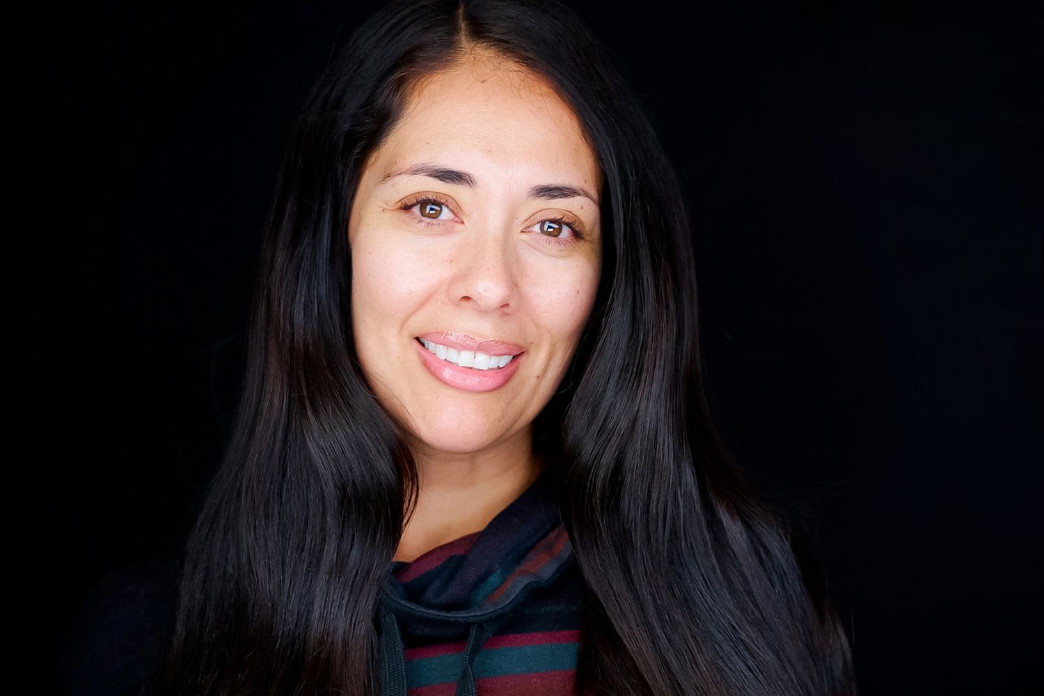Smiling woman with long dark hair wearing a striped cowl neck top, posed against a solid black background in a studio portrait.