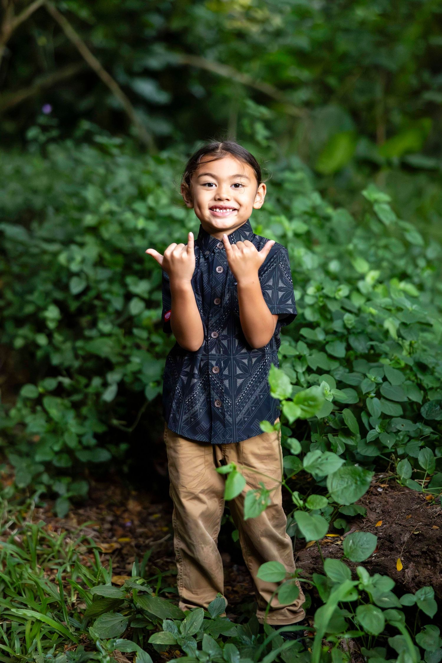 Smiling young boy in a dark patterned shirt and khaki pants makes shaka hand gestures while standing among lush green plants outdoors.