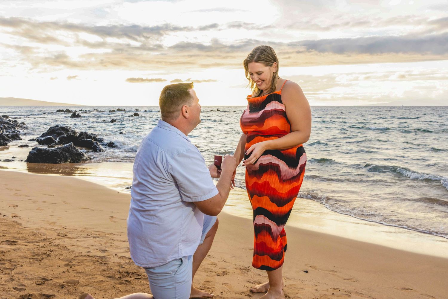 Man in a light blue shirt kneels on one knee on a sandy beach proposing to a smiling woman in a colorful dress at sunset. Wailea