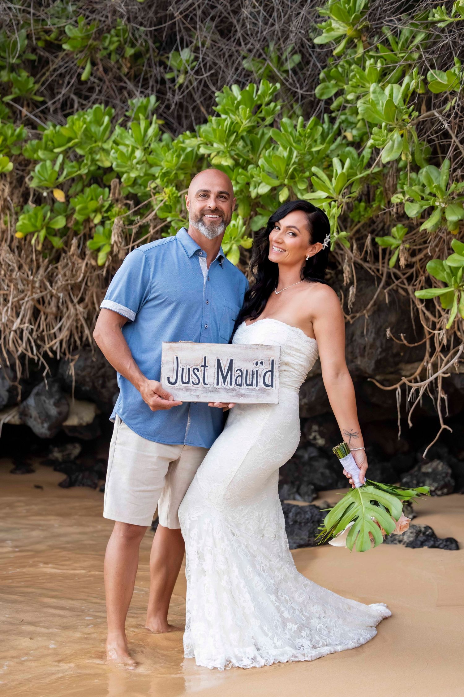 Newlywed couple in a white wedding dress and blue shirt hold a Just Maui'd sign while standing barefoot on a Maui beach. Makena