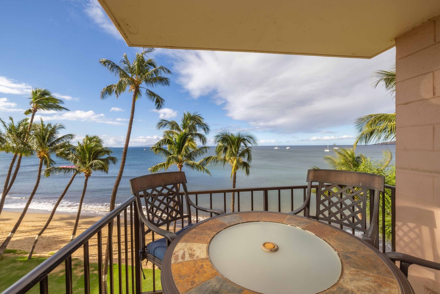 Oceanfront condo balcony with a mosaic table and two chairs overlooking swaying palm trees, a sandy beach, and calm blue tropical waters.