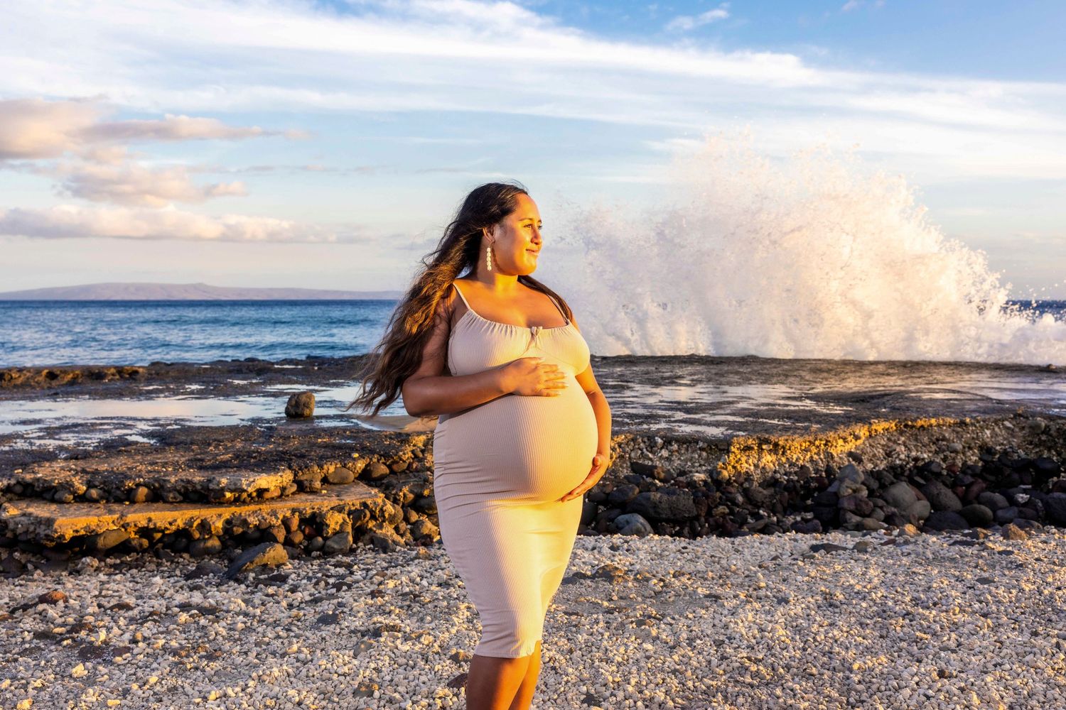 Pregnant woman in a fitted cream dress cradling her belly on a rocky beach as a large ocean wave crashes behind her. Olowalu
