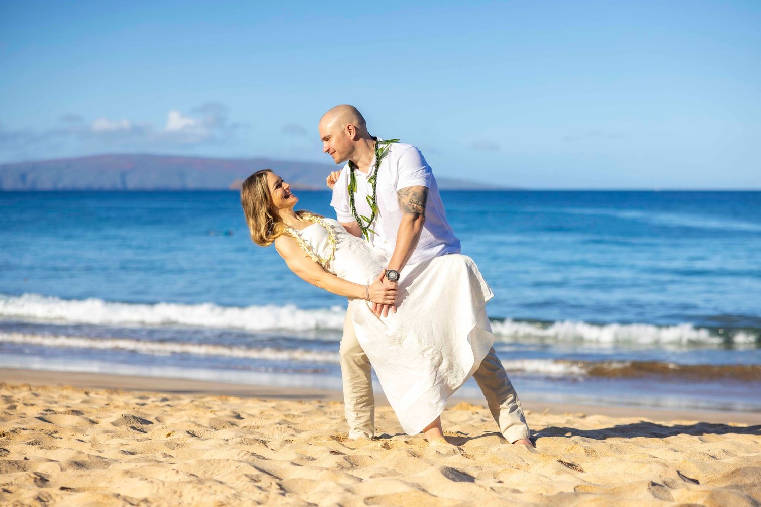 Groom dips bride in a romantic pose on a sunny Maui beach during a Hawaiian wedding, both wearing white and leis. Wailea