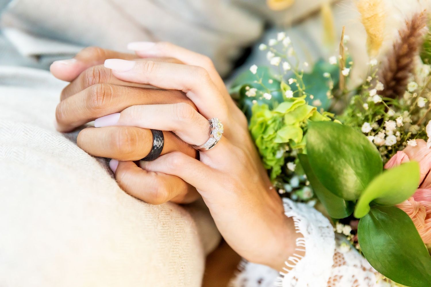 Close-up of a bride and groom holding hands with wedding rings, beside a green and white floral bridal bouquet. Lahaina