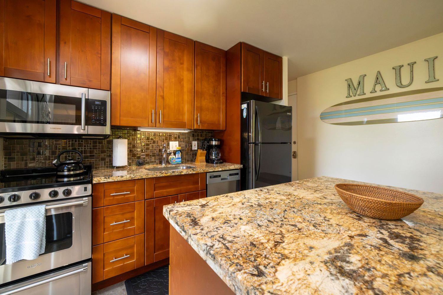 Updated kitchen with warm wood cabinets, granite island with wooden bowl, stainless steel appliances and a MAUI wall sign.