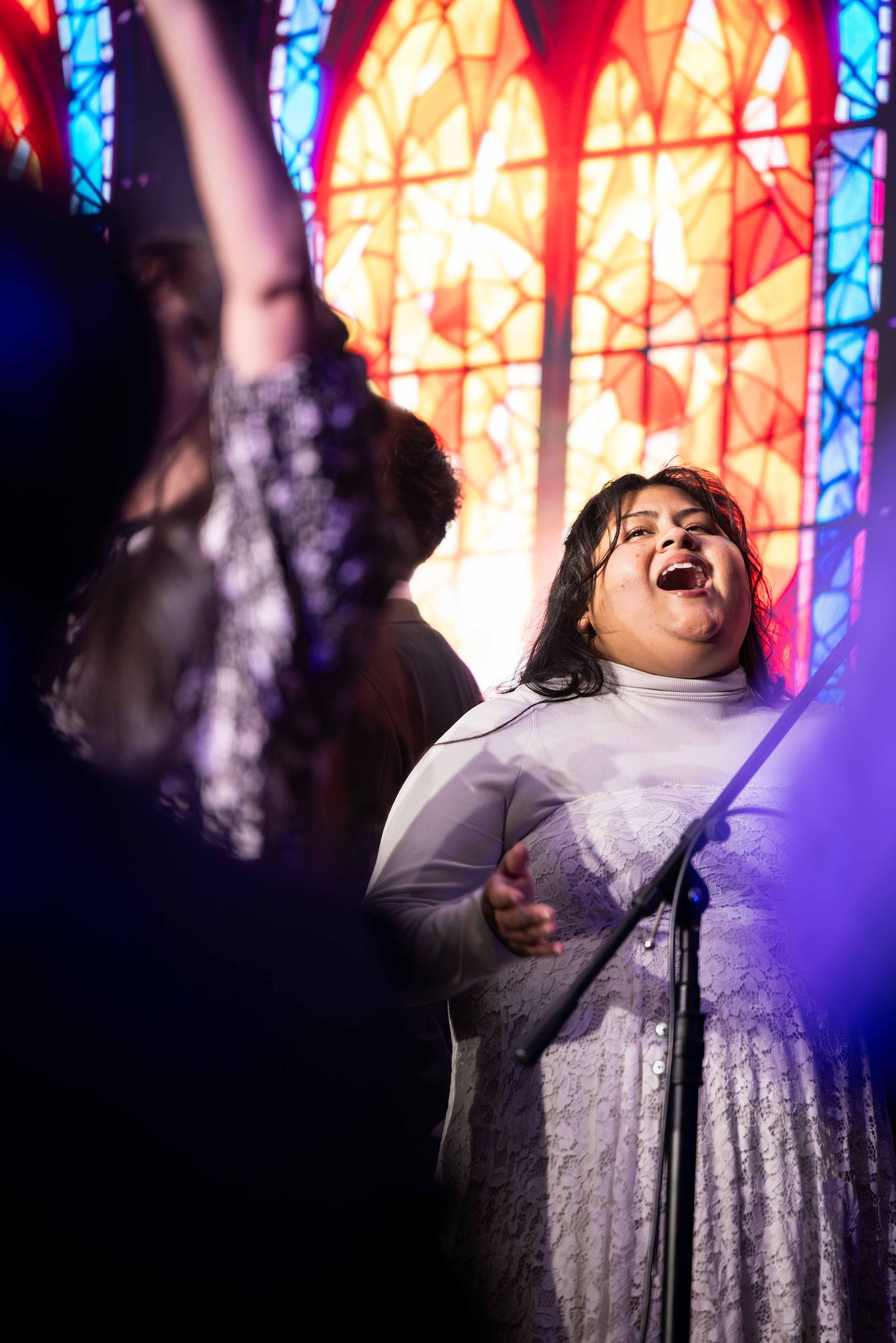 Woman in a white lace dress sings passionately into a microphone in front of a glowing stained glass window as another person raises an arm.