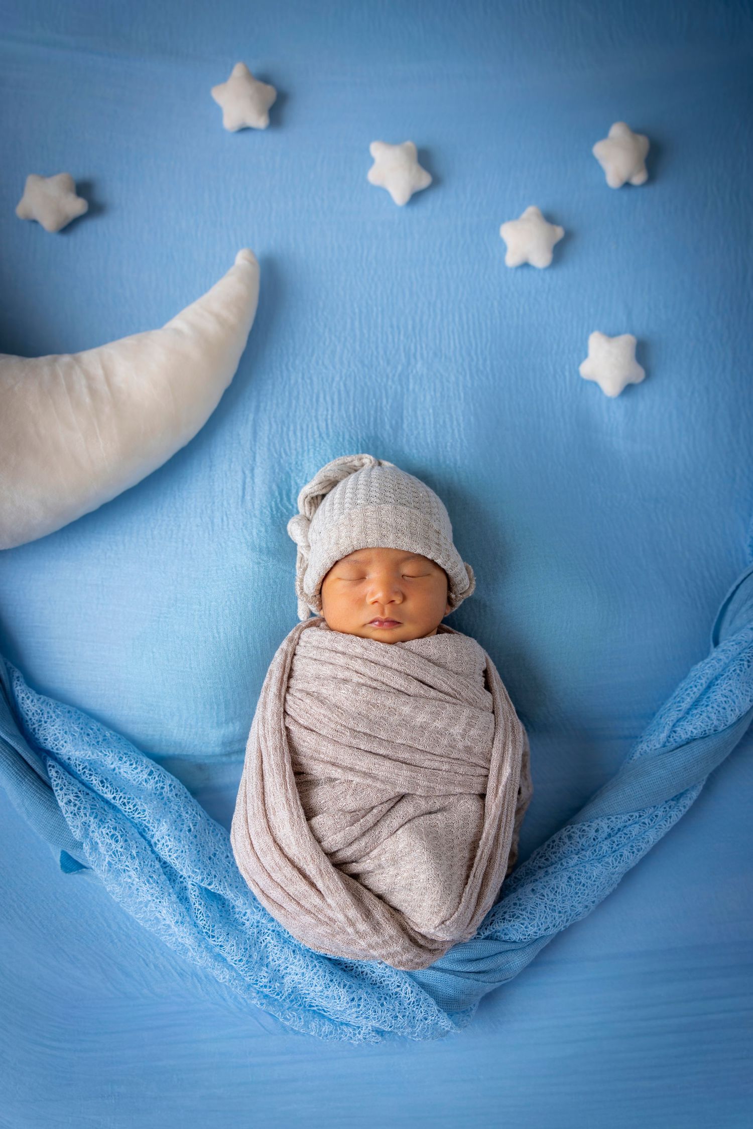 Sleeping newborn swaddled in gray wrap and knit cap on a blue backdrop with a plush crescent moon and felt stars.