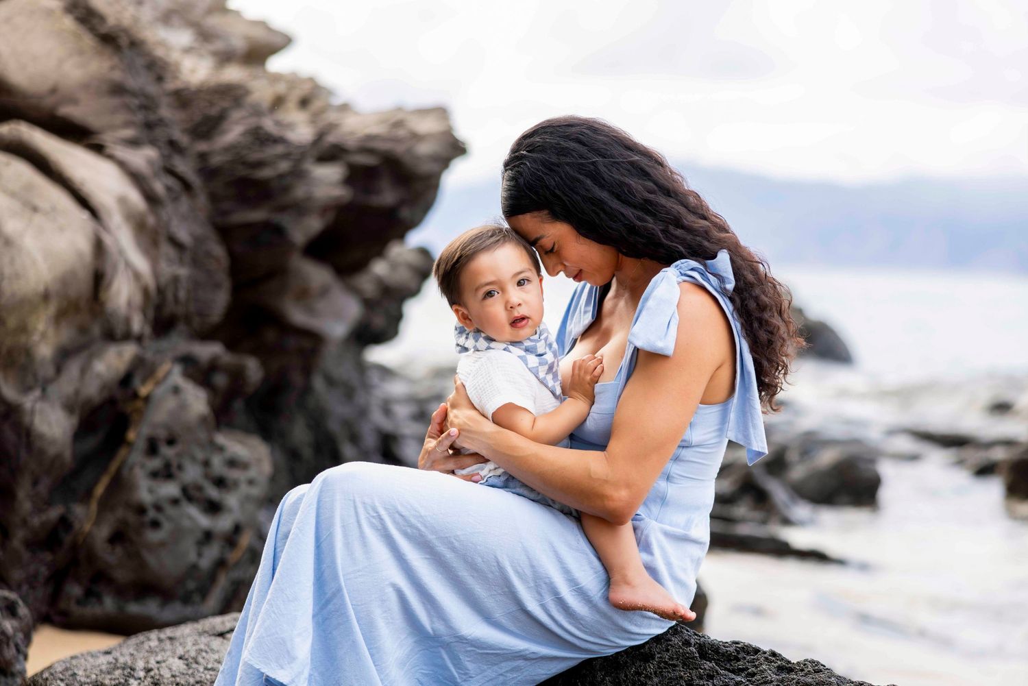 Mother in a flowing light blue dress sits on coastal rocks holding her smiling toddler, with the ocean and a hazy shoreline behind them.