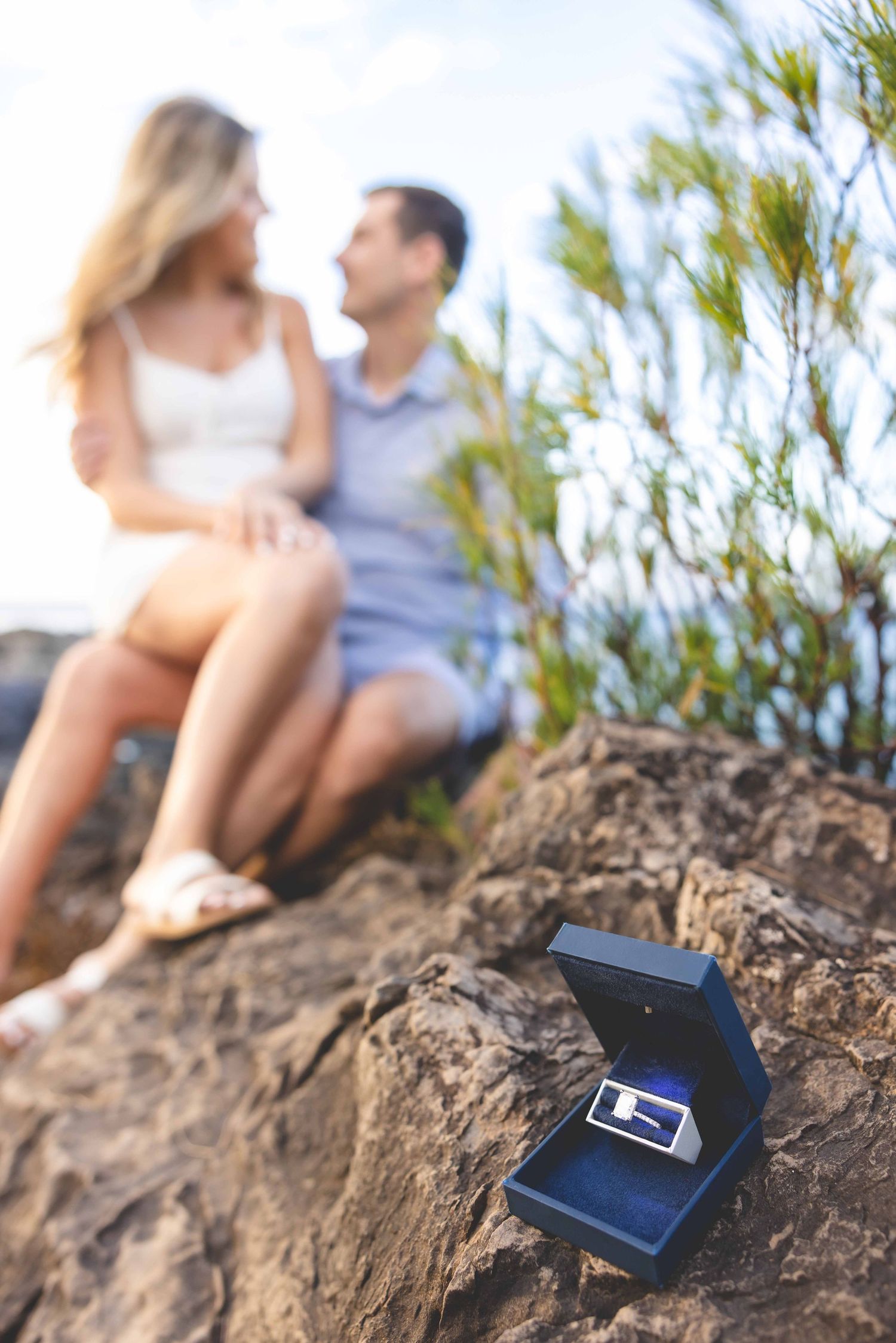 Open ring box with a diamond ring on a rocky ledge, with a couple sitting together blurred in the background during a proposal. Kapalua Bay