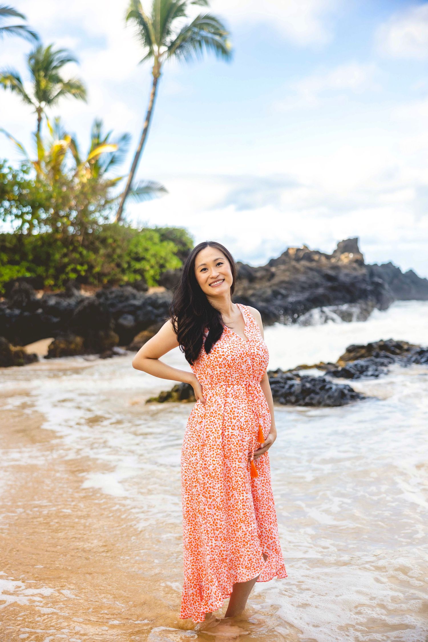 Woman in a coral floral maxi dress stands barefoot in shallow ocean water on a tropical beach with lava rocks and palm trees. Makena