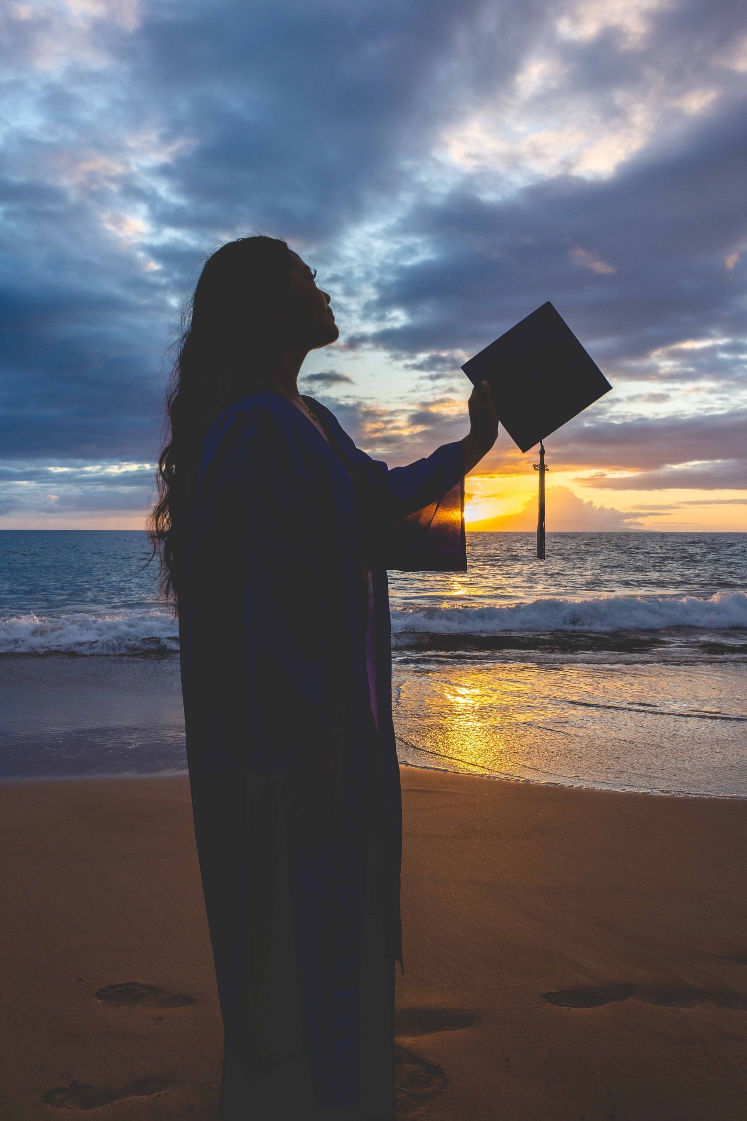 Silhouette of a graduate in a gown holding a mortarboard cap up toward the sky on a beach at sunset, with waves and dramatic clouds. Kihei